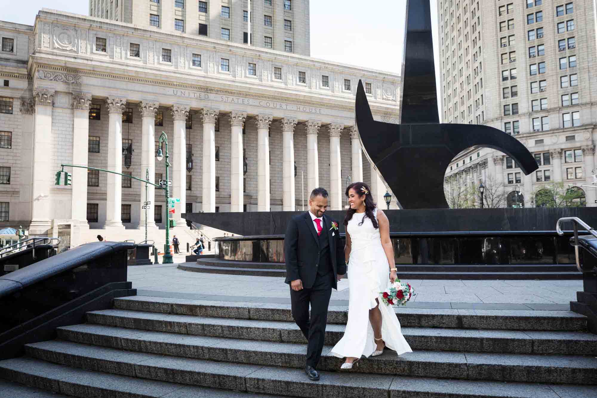 Bride and groom walking down steps in front of sword statue and large building with columns  at a NYC City Hall wedding