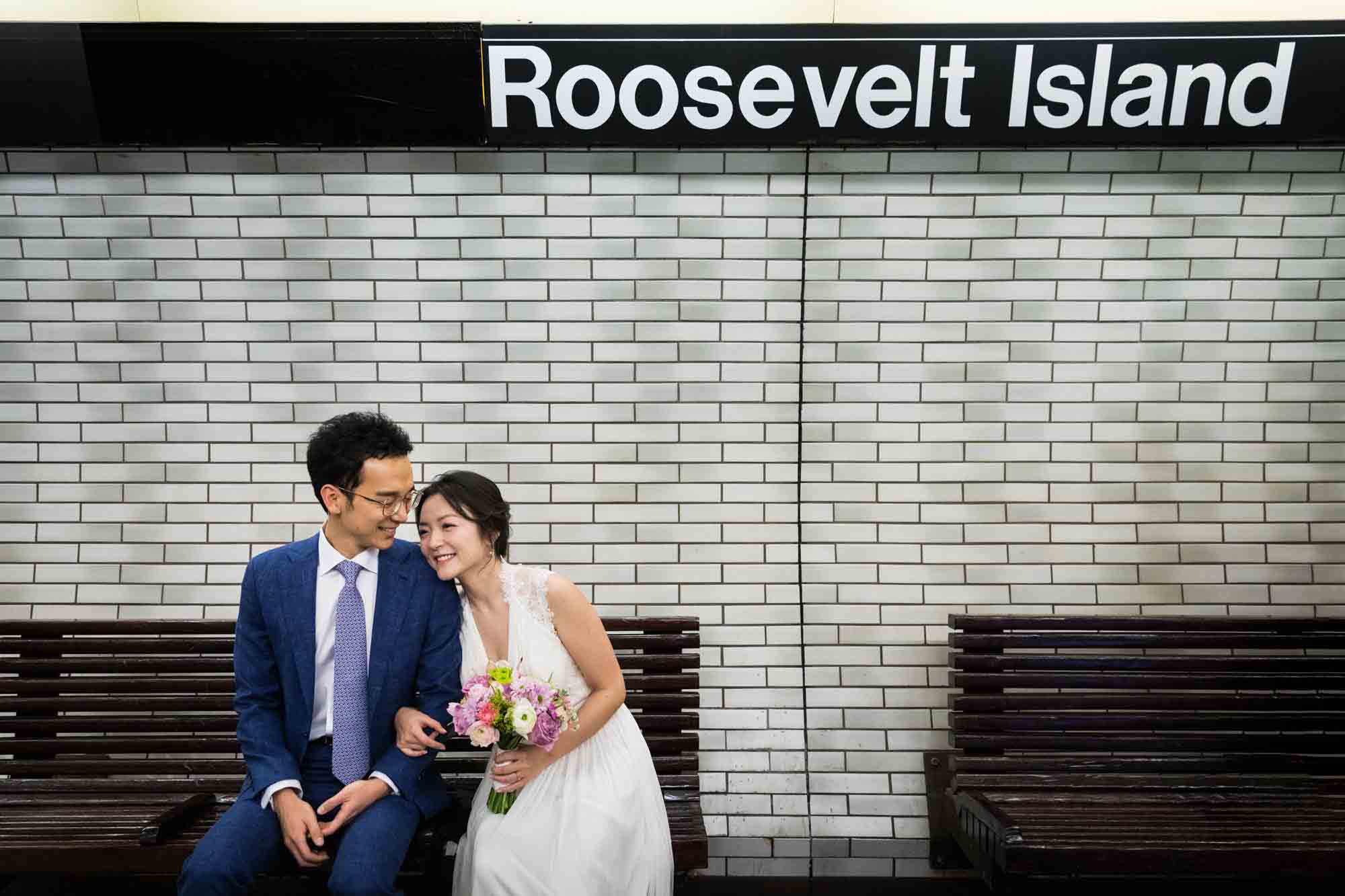 Couple sitting on bench in subway station under Roosevelt Island sign during a Roosevelt Island engagement photo shoot