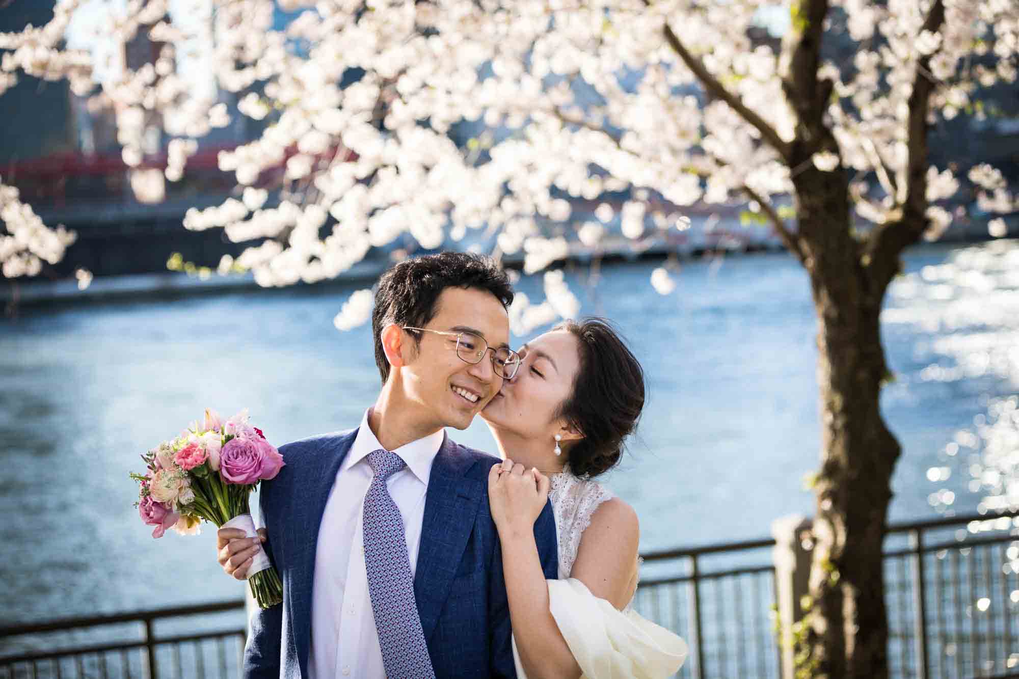 Couple kissing in front of cherry blossom trees and waterfront during a Roosevelt Island engagement photo shoot