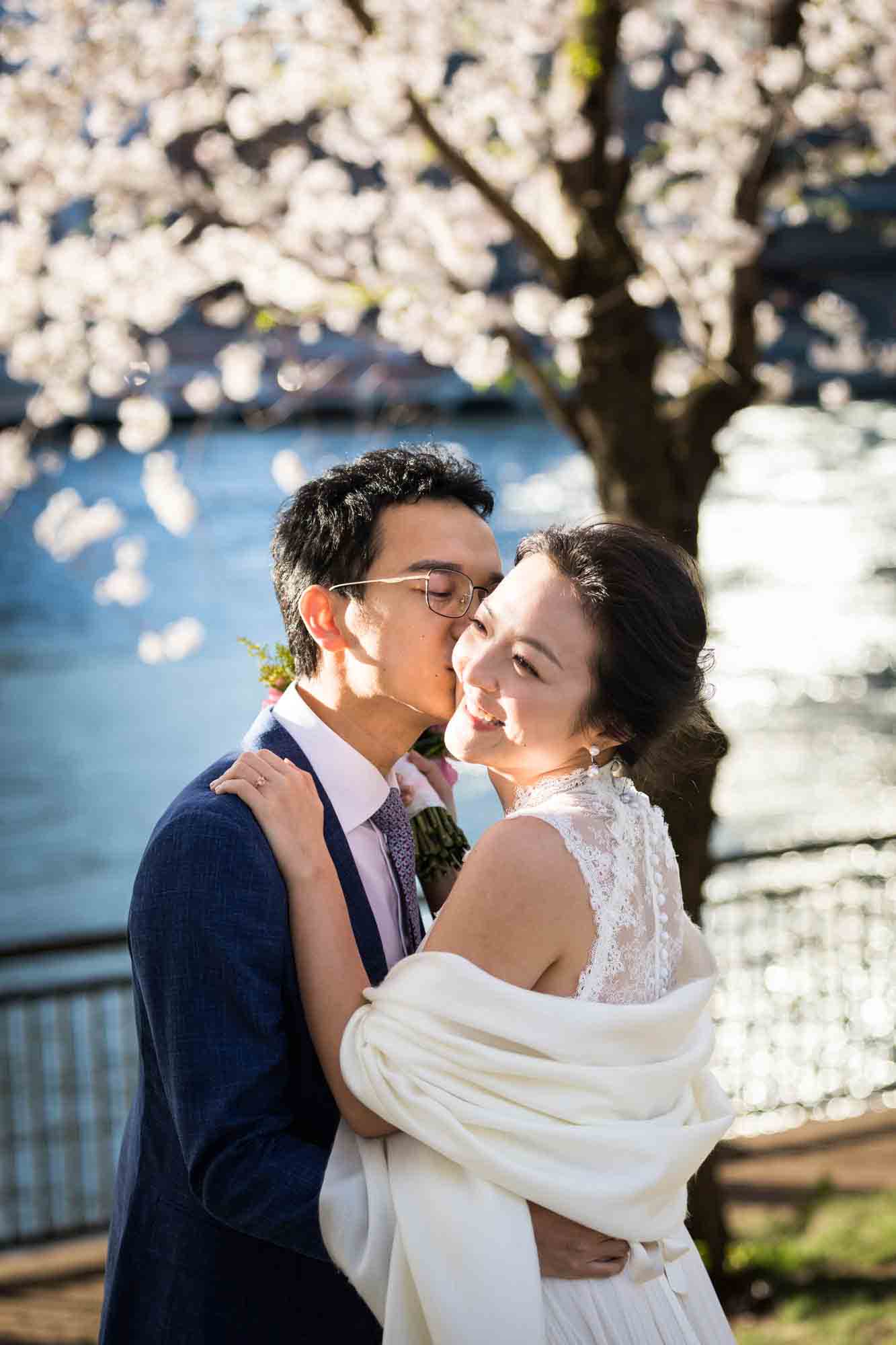 Couple kissing in front of cherry blossom trees and waterfront during a Roosevelt Island engagement photo shoot
