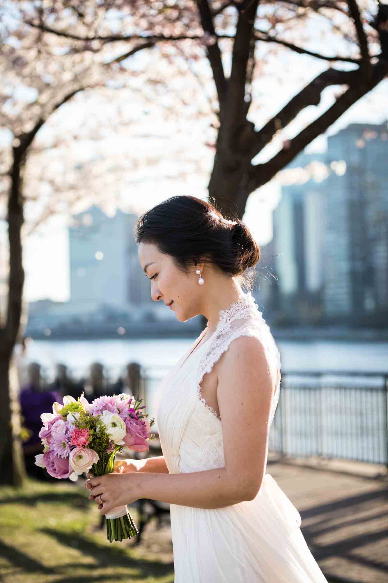 Woman wearing white dress looking down at flower bouquet Couple walking under cherry blossom trees during a Roosevelt Island engagement photo shoot