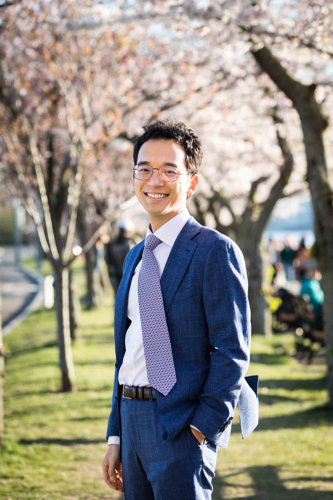 Man wearing blue suit and blue tie in front of cherry blossom trees Couple walking under cherry blossom trees during a Roosevelt Island engagement photo shoot