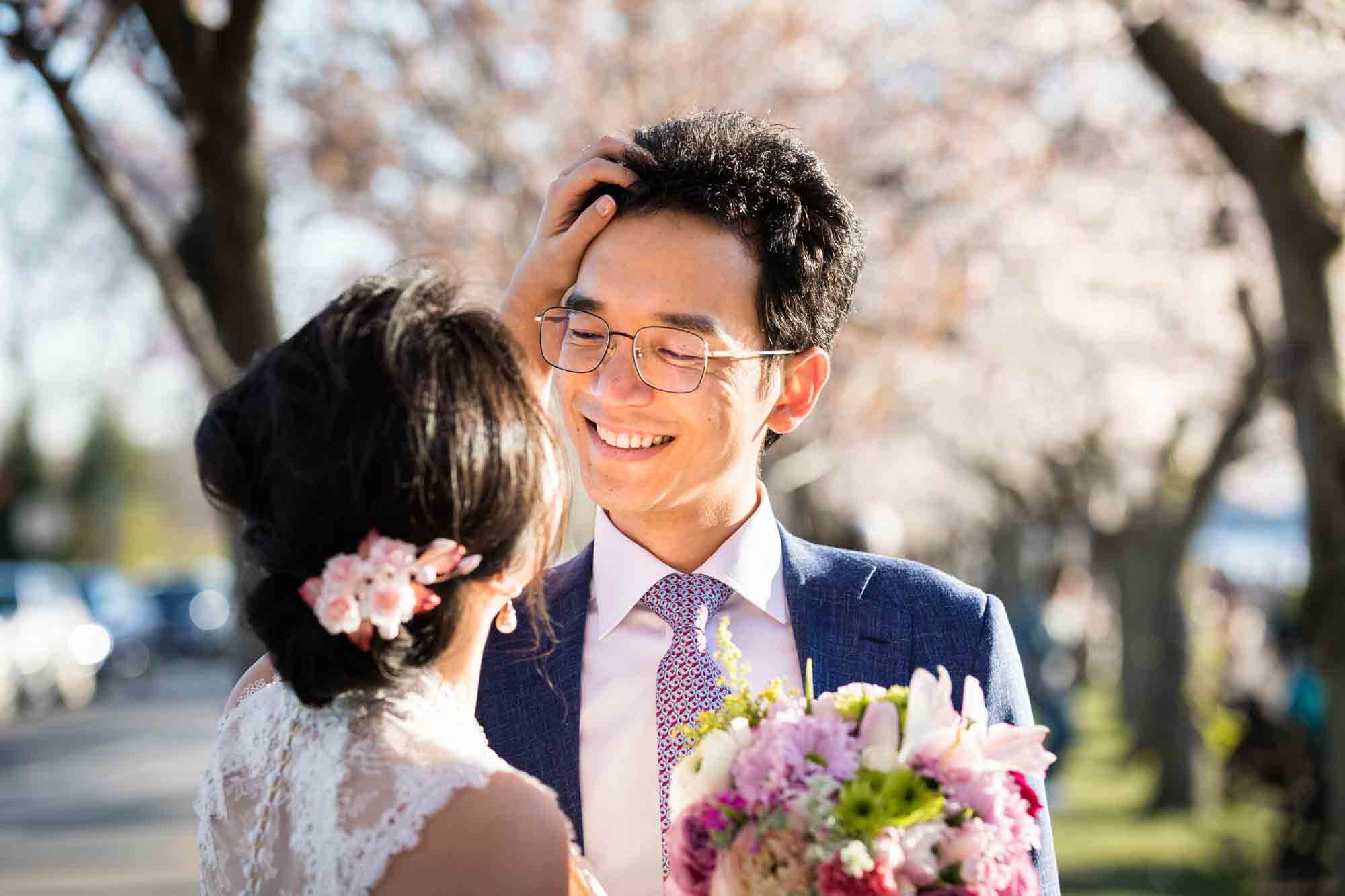 Woman holding flowers and touching man's hair Couple walking under cherry blossom trees during a Roosevelt Island engagement photo shoot