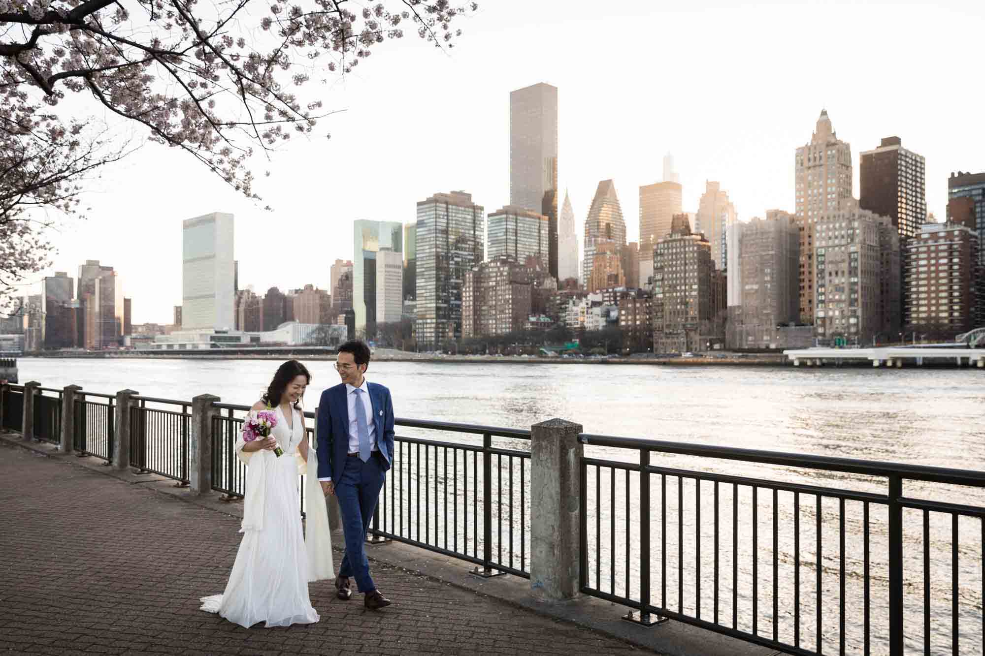Roosevelt Island engagement photos of couple walking along pathway in front of waterfront and NYC skyline