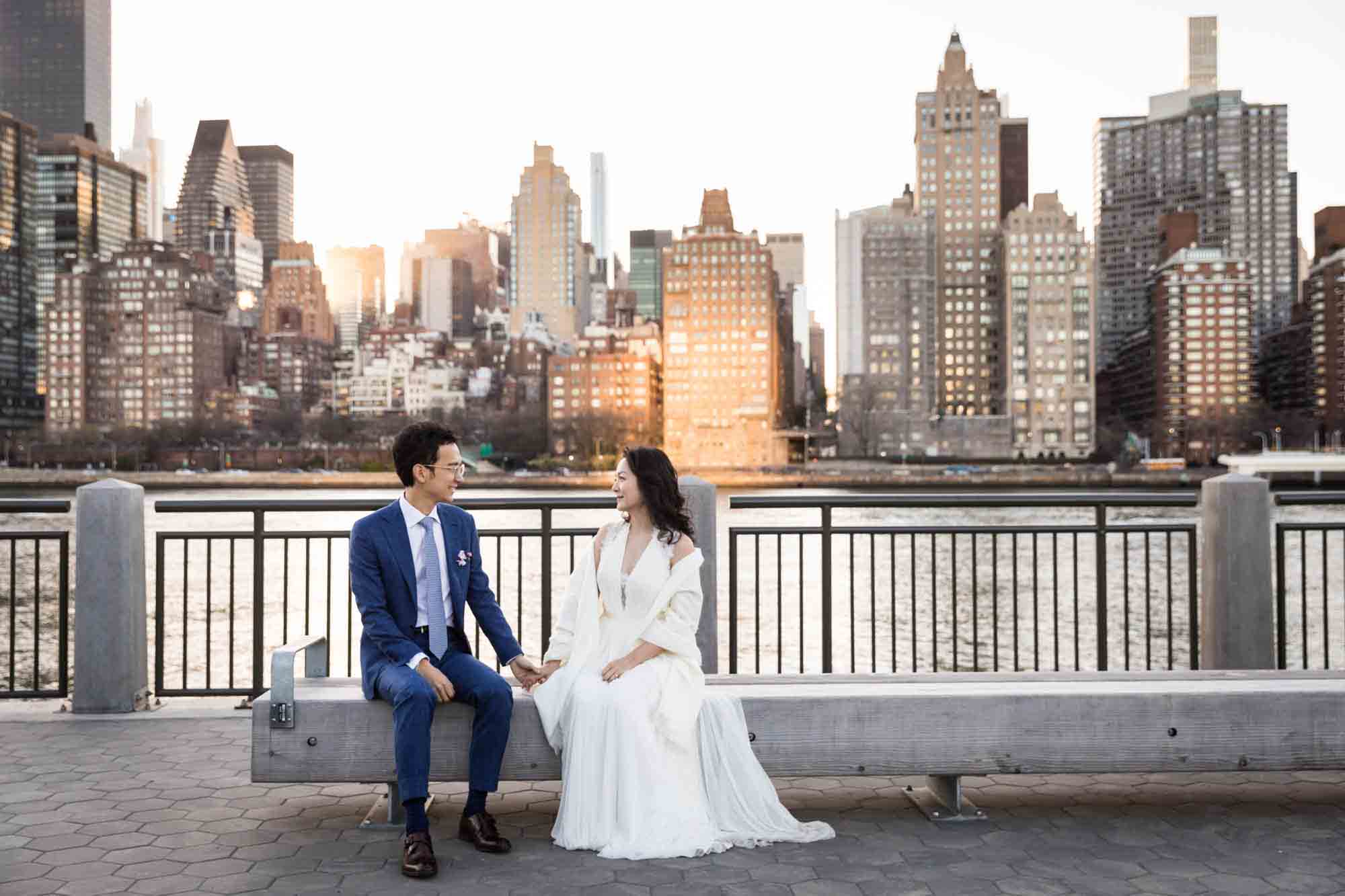 Roosevelt Island engagement photos of couple sitting on bench holding hands in front of NYC skyline