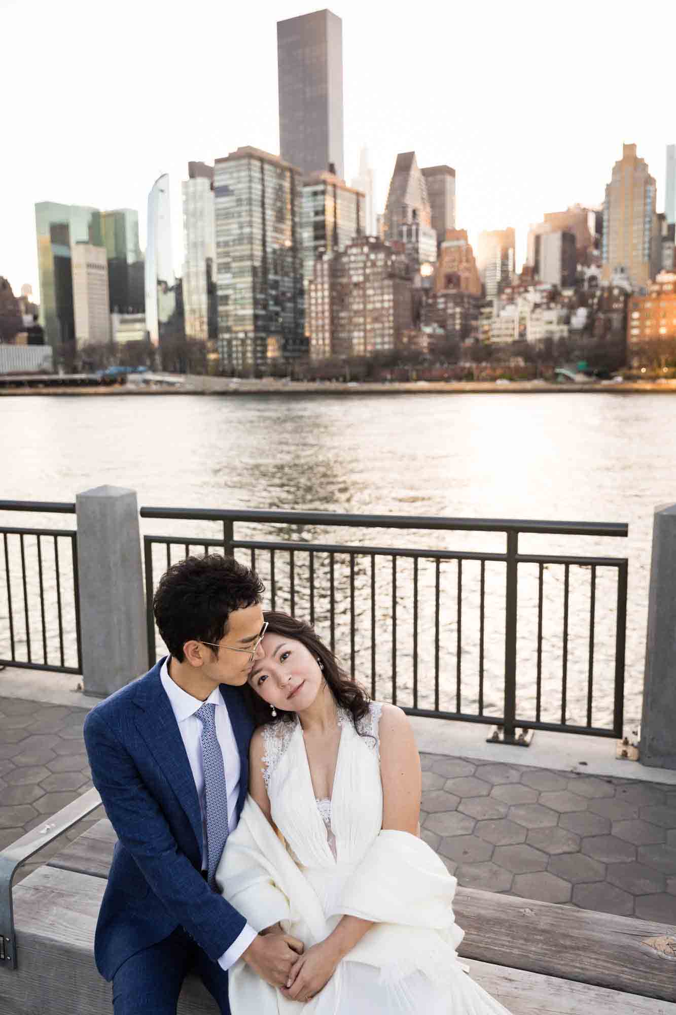 Roosevelt Island engagement photos of couple sitting on bench hugging in front of NYC skyline