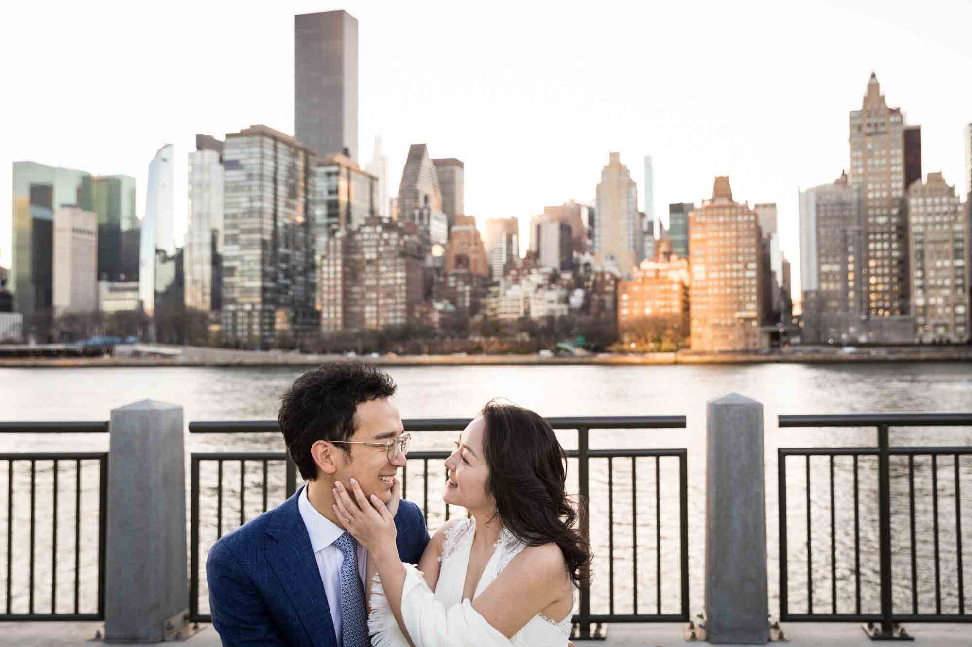 Roosevelt Island engagement photos of couple sitting on bench with woman touching man's face in front of NYC skyline