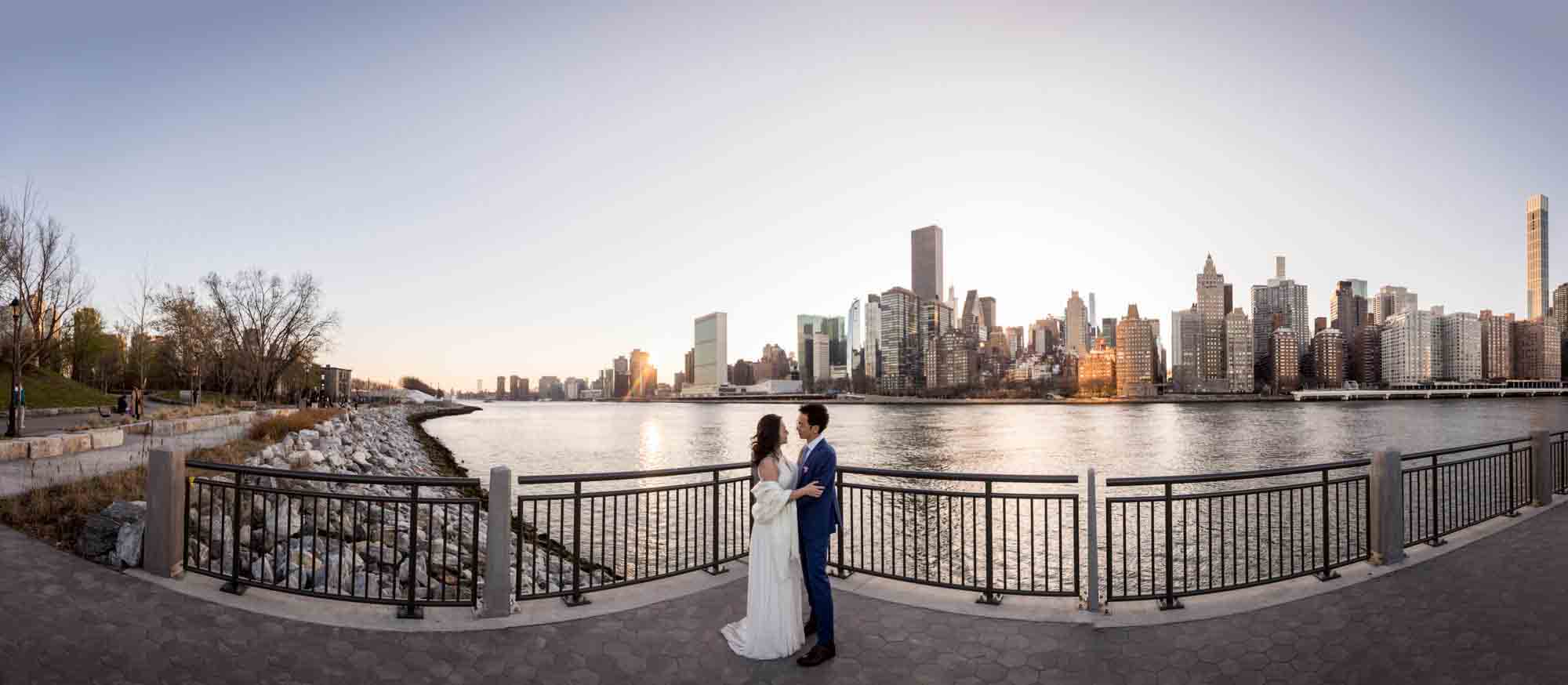 Panorama of couple on standing beside waterfront in front of NYC skyline during a Roosevelt Island engagement photo shoot