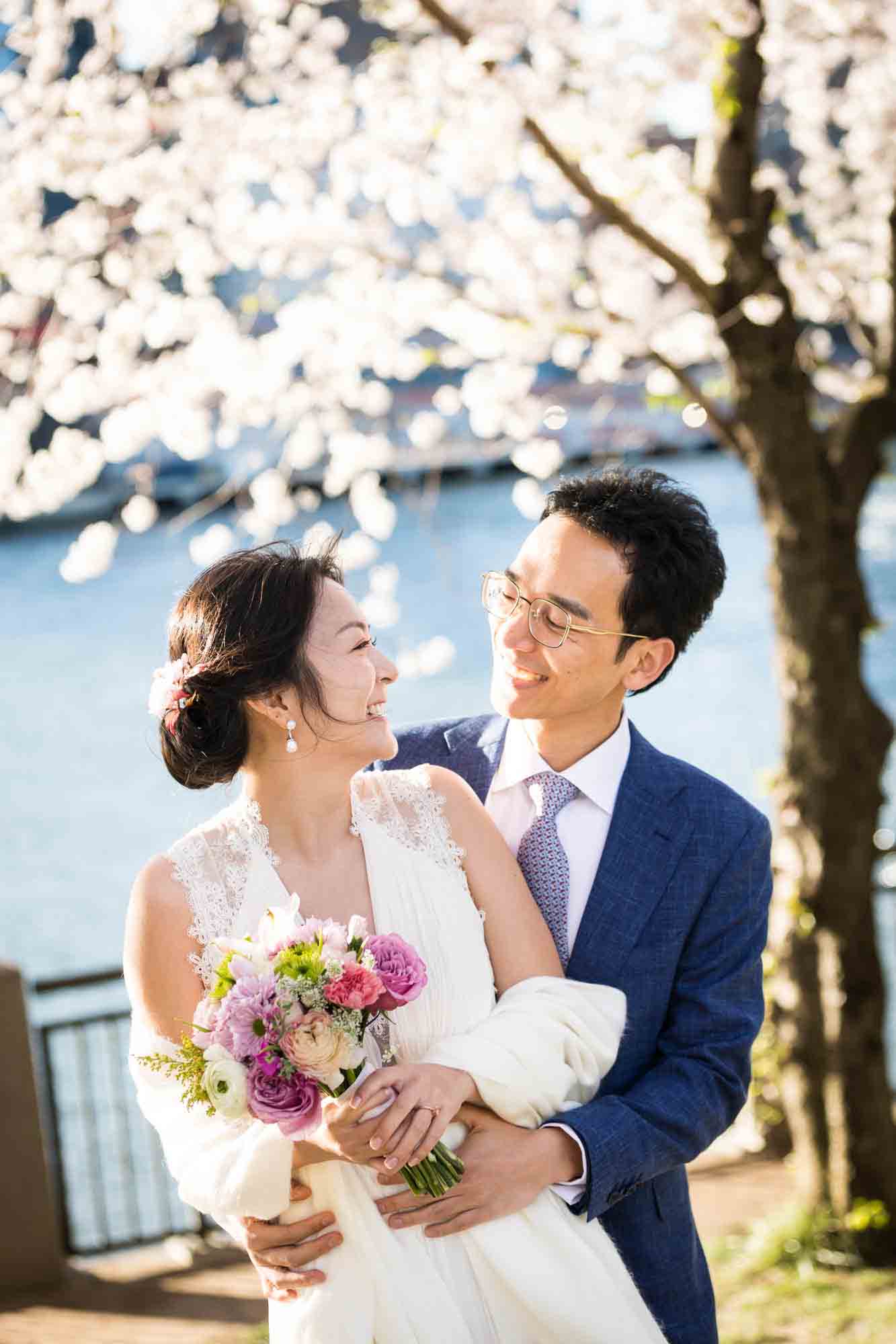 Couple hugging in front of cherry blossom trees and waterfront during a Roosevelt Island engagement photo shoot
