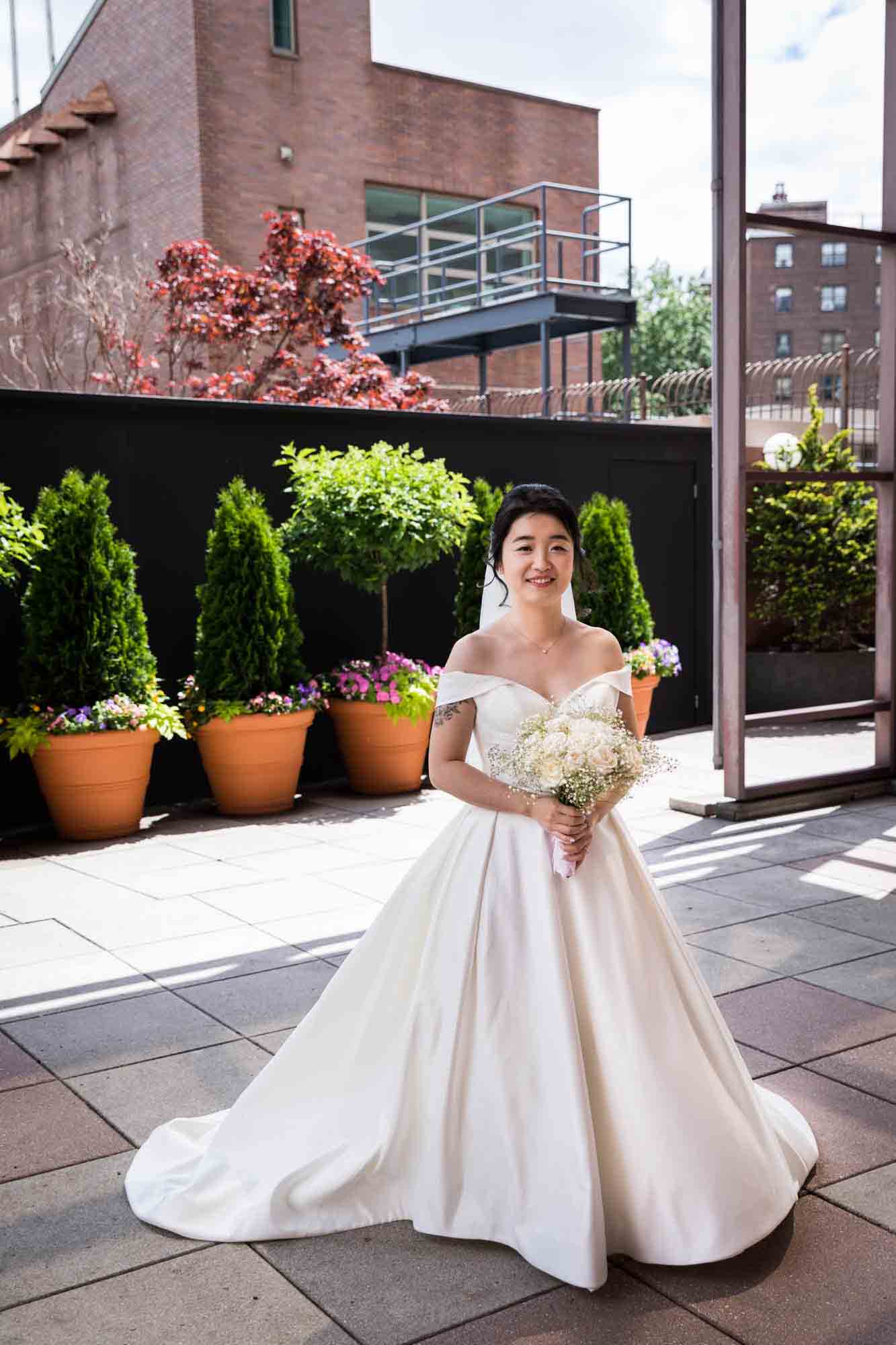 Bride standing in front of potted bushes in long off-shoulder white dress and holding flower bouquet at a Sheraton LaGuardia East Hotel wedding