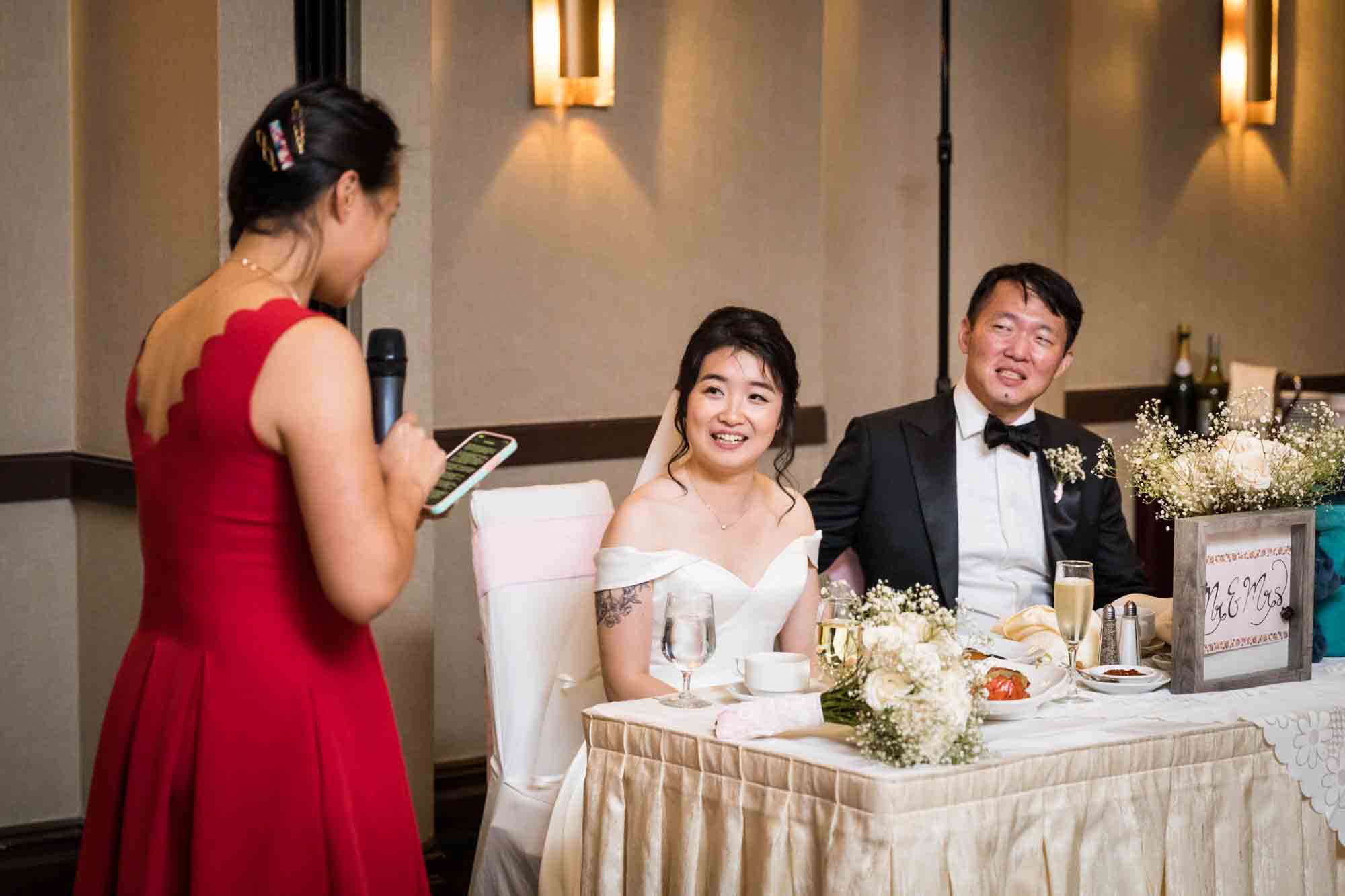 Bride and groom sitting at table listening to woman in red dress talking into microphone at a Sheraton LaGuardia East Hotel wedding