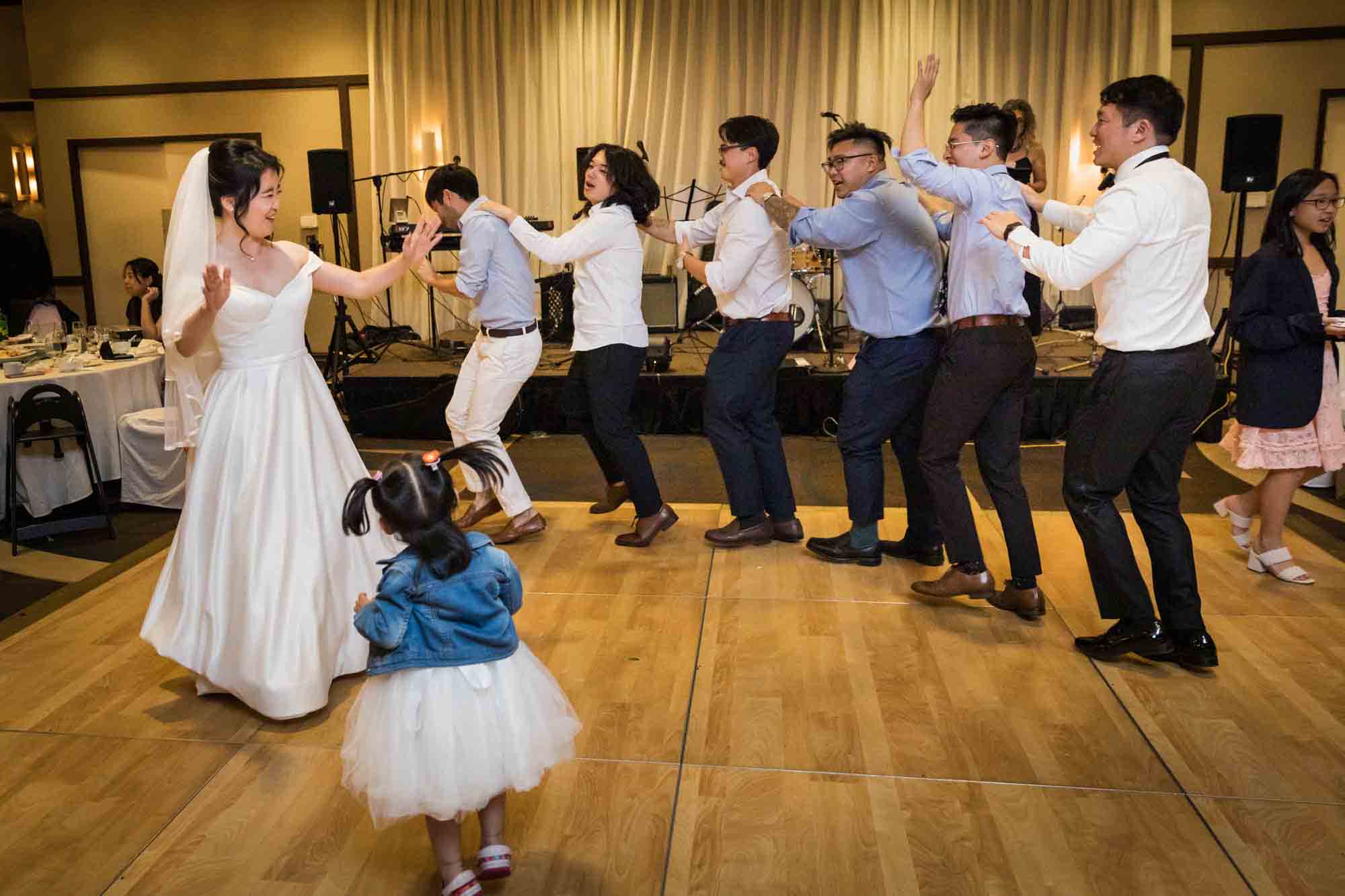 Bride and little girl dancing in front of six men dancing in a line at a Sheraton LaGuardia East Hotel wedding