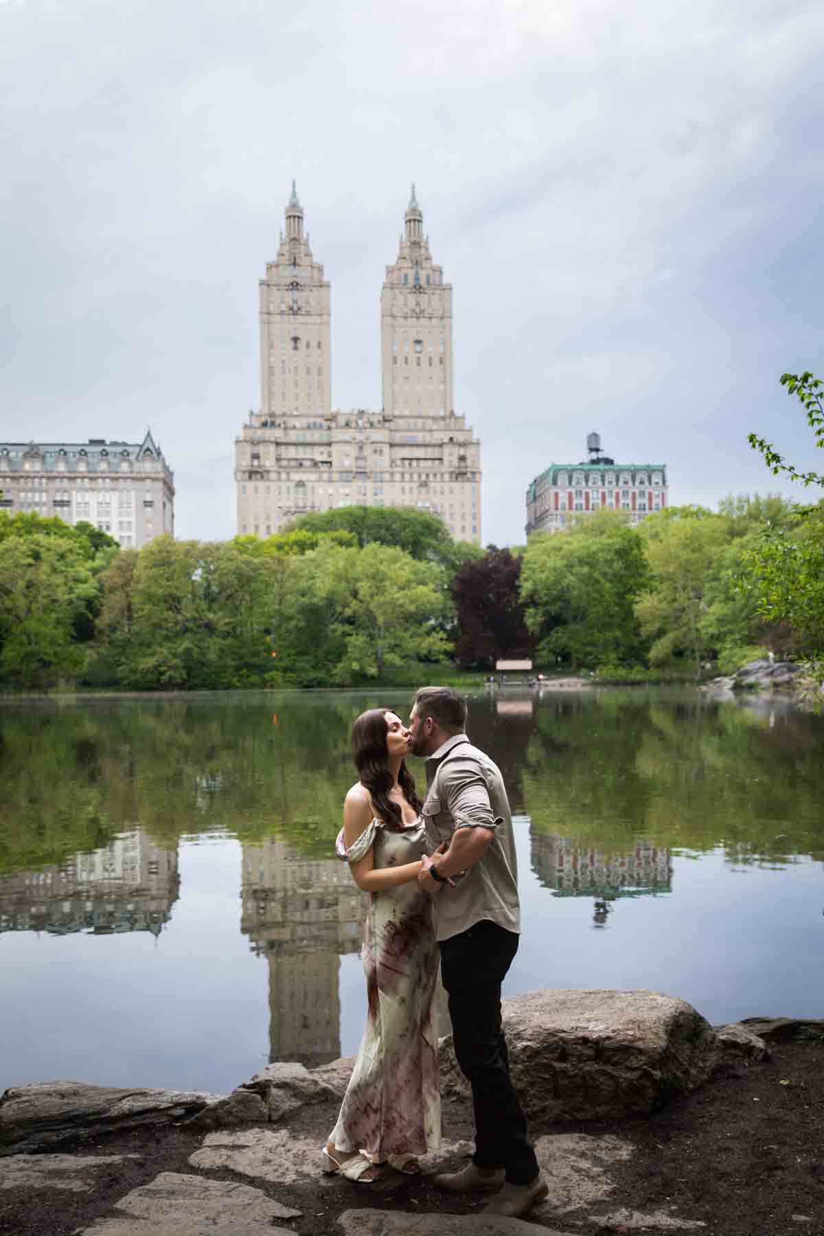 Couple kissing in front of lake with two tall buildings in background during a Central Park surprise proposal