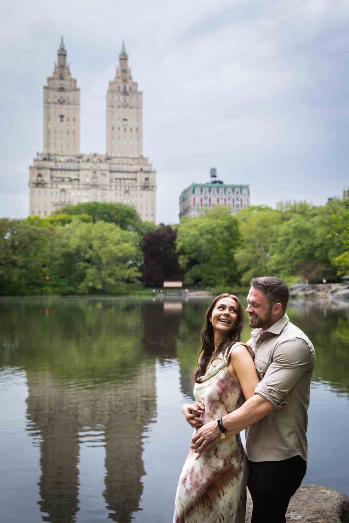 Couple hugging in front of lake with two tall buildings in background during a Central Park surprise proposal