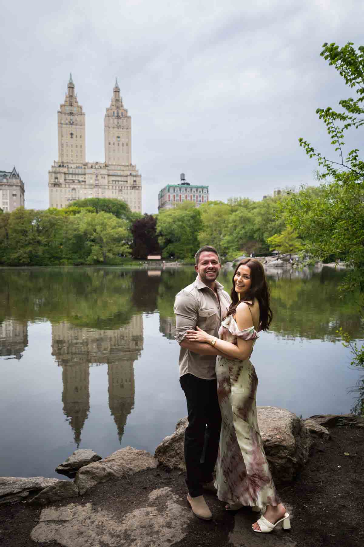 Couple hugging in front of lake with two tall buildings in background during a Central Park surprise proposal