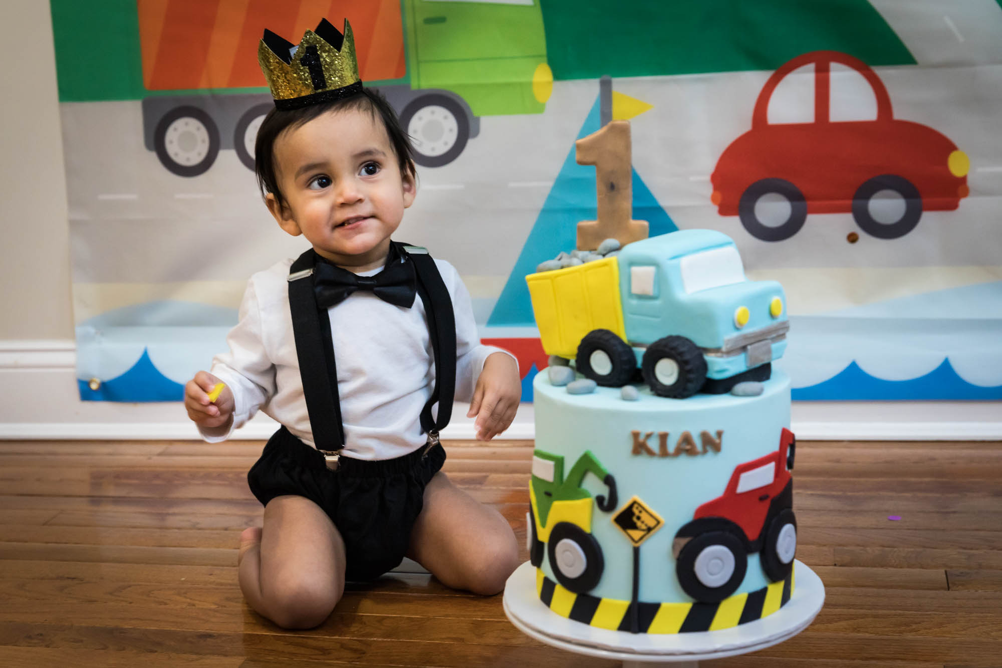 Little boy sitting on wooden floor in front of truck-themed cake wearing gold crown and black bow tie for an article on cake smash tips