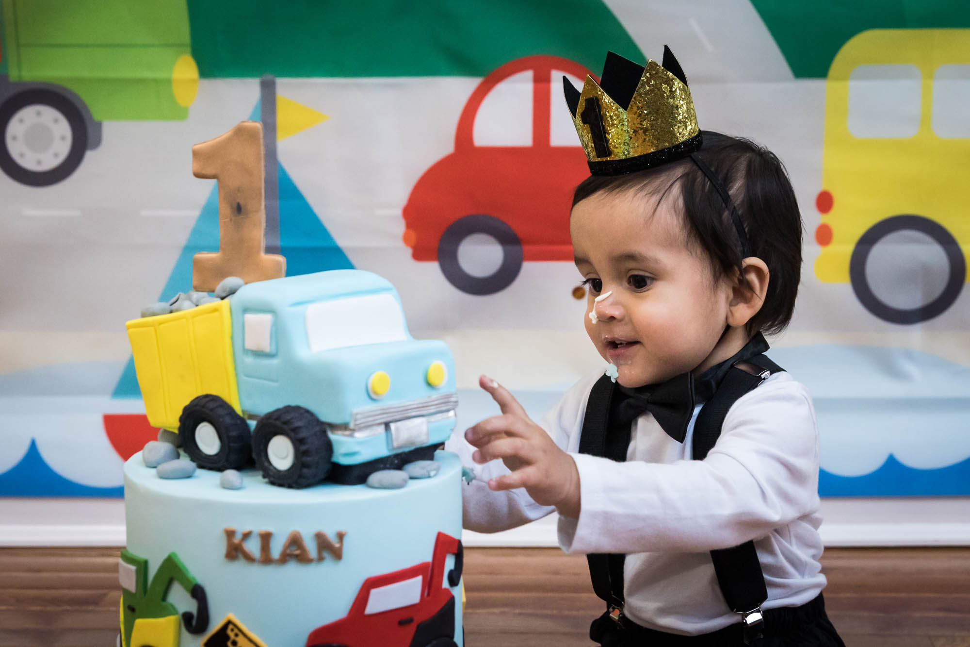 Little boy sitting on wooden floor touching truck-themed cake wearing gold crown and black bow tie for an article on cake smash tips