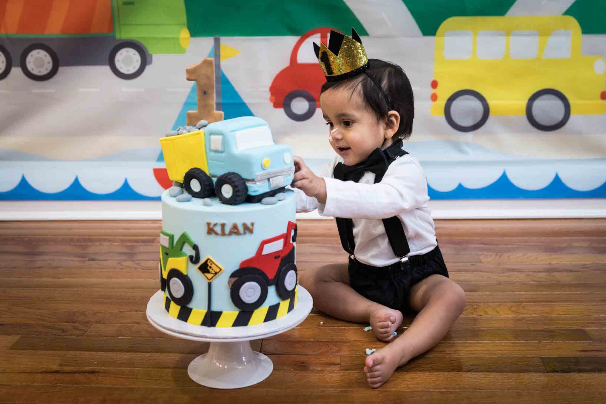 Little boy sitting on wooden floor touching truck-themed cake wearing gold crown and black bow tie for an article on cake smash tips