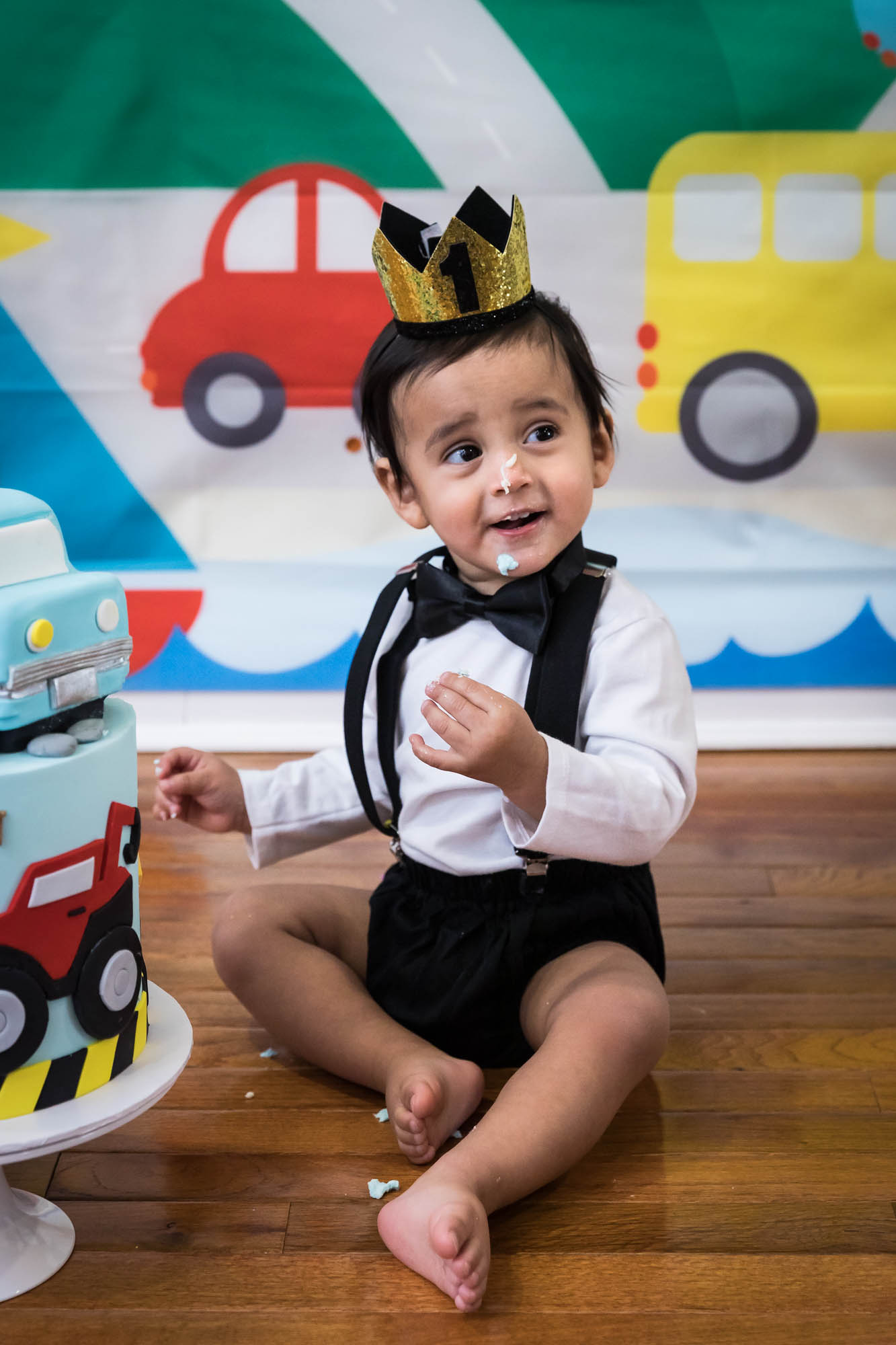 Little boy sitting on wooden floor in front of cake wearing gold crown and black bow tie for an article on cake smash tips