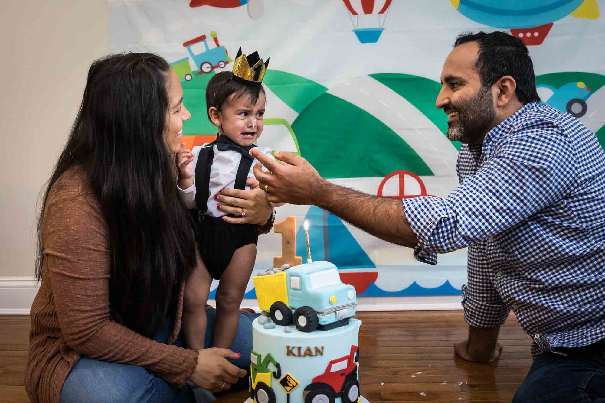 Parents holding crying little boy wearing gold crown with father showing finger of frosting to little boy for an article on cake smash tips