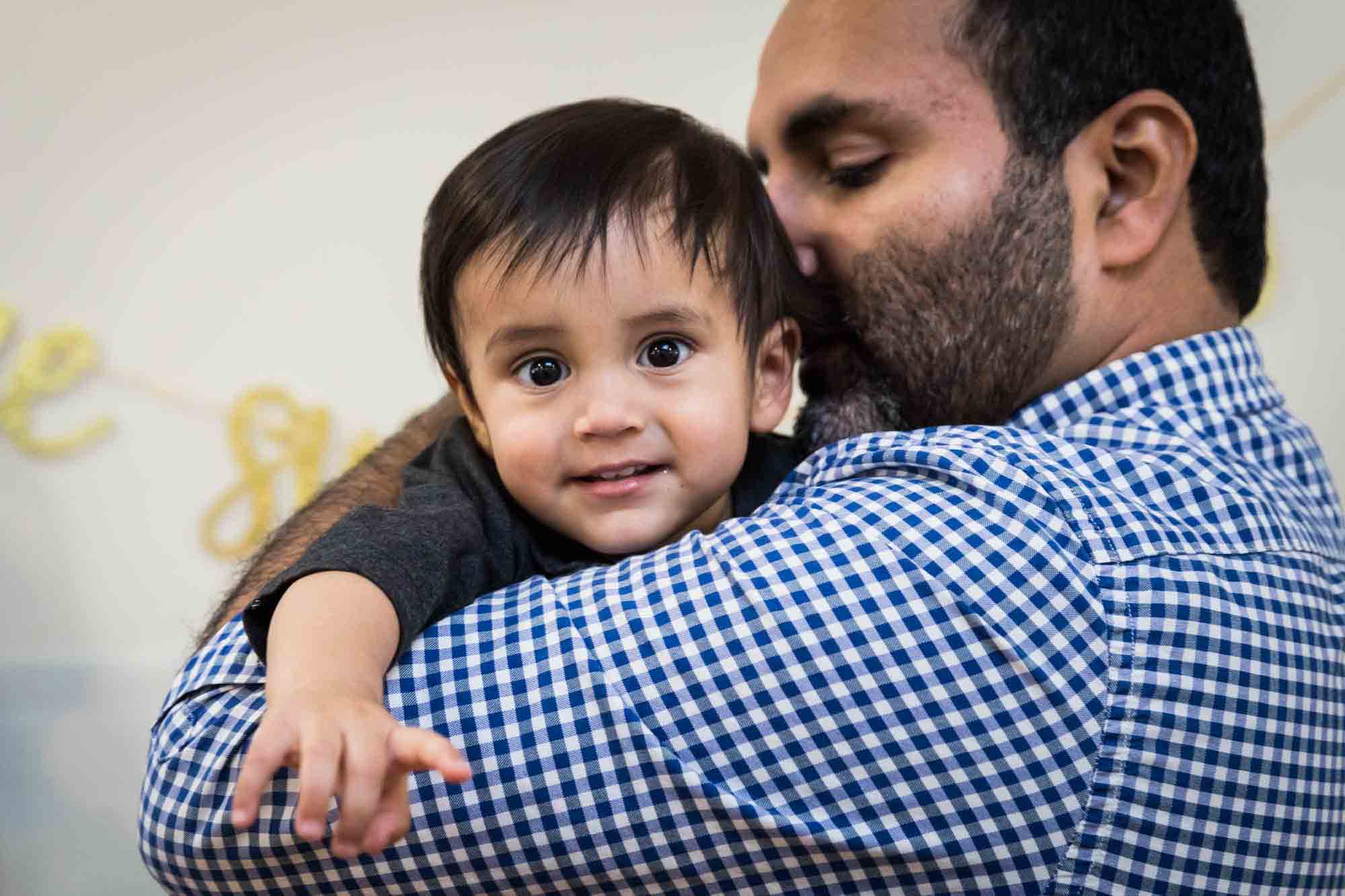 Father wearing blue and white checkered shirt kissing little boy on the head while holding him for an article on cake smash tips