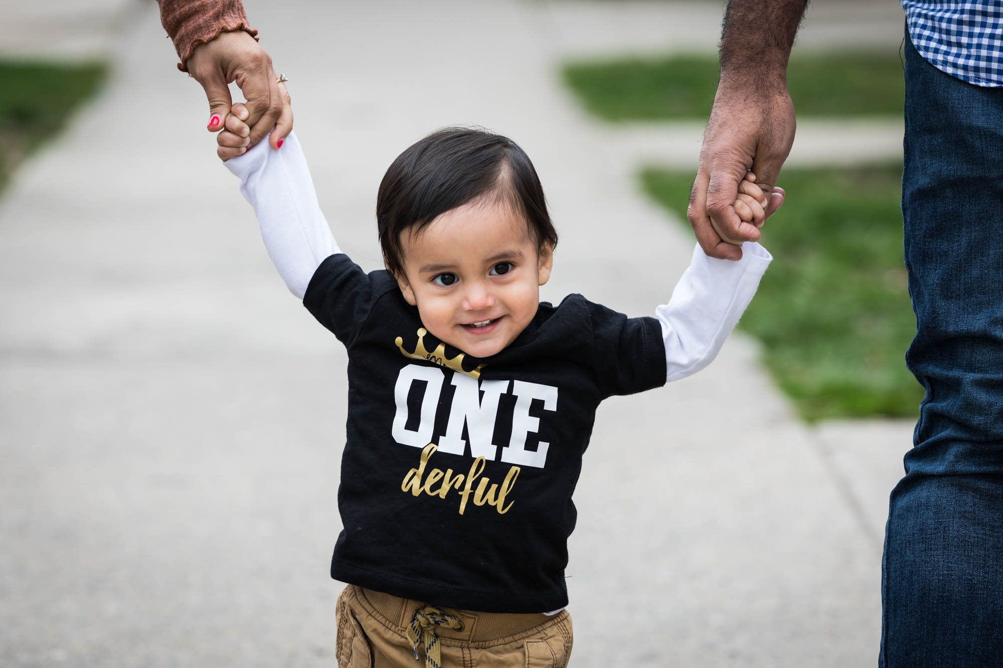 Little boy standing on sidewalk holding hands of male and female adult wearing black shirt that says 'Onederful'