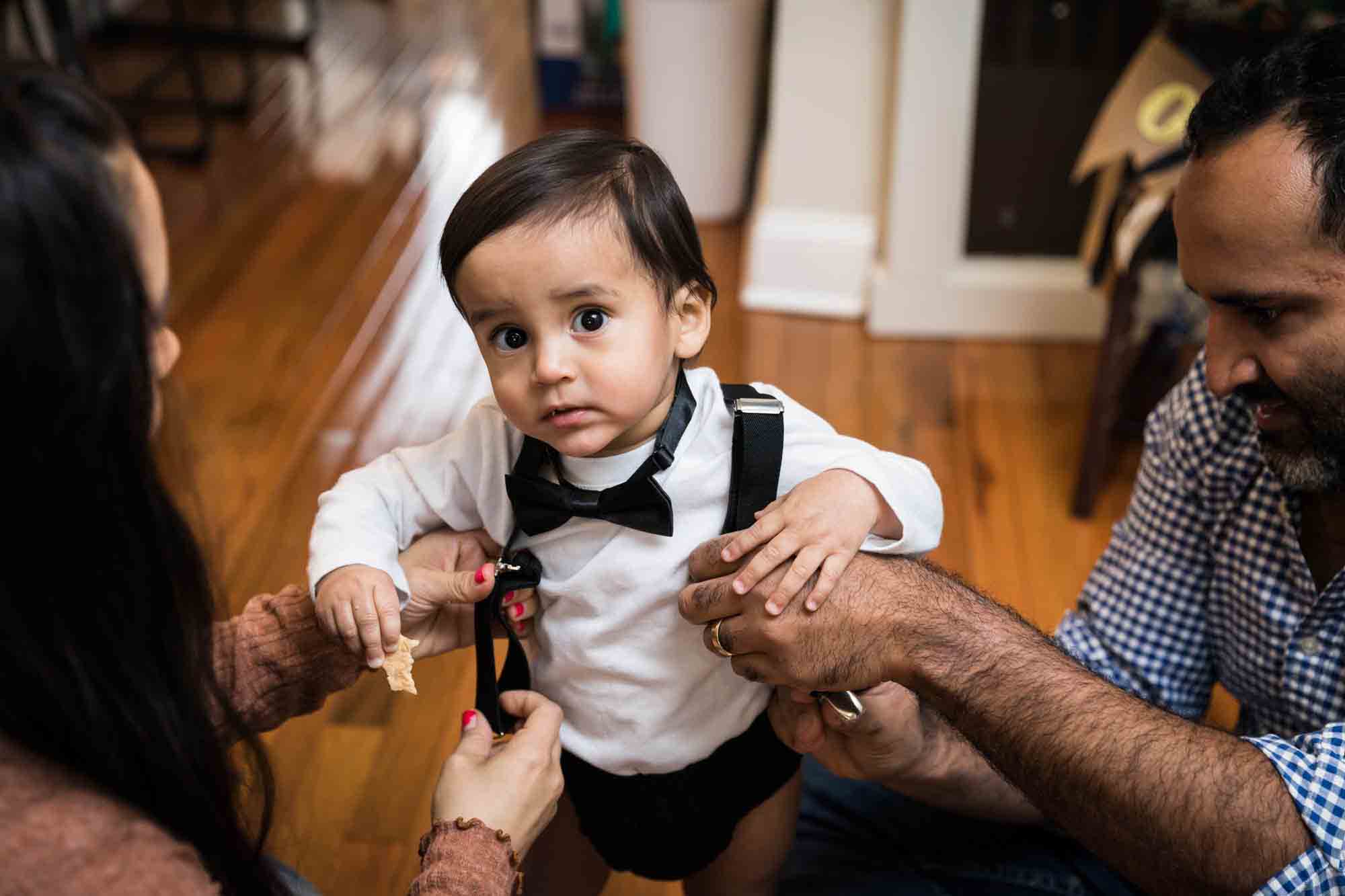 Little boy standing on wooden floor with mother and father adjusting his black suspenders