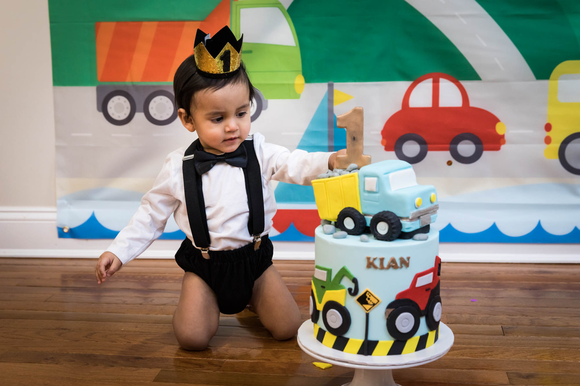 Little boy sitting on wooden floor touching truck-themed cake wearing gold crown and black bow tie for an article on cake smash tips