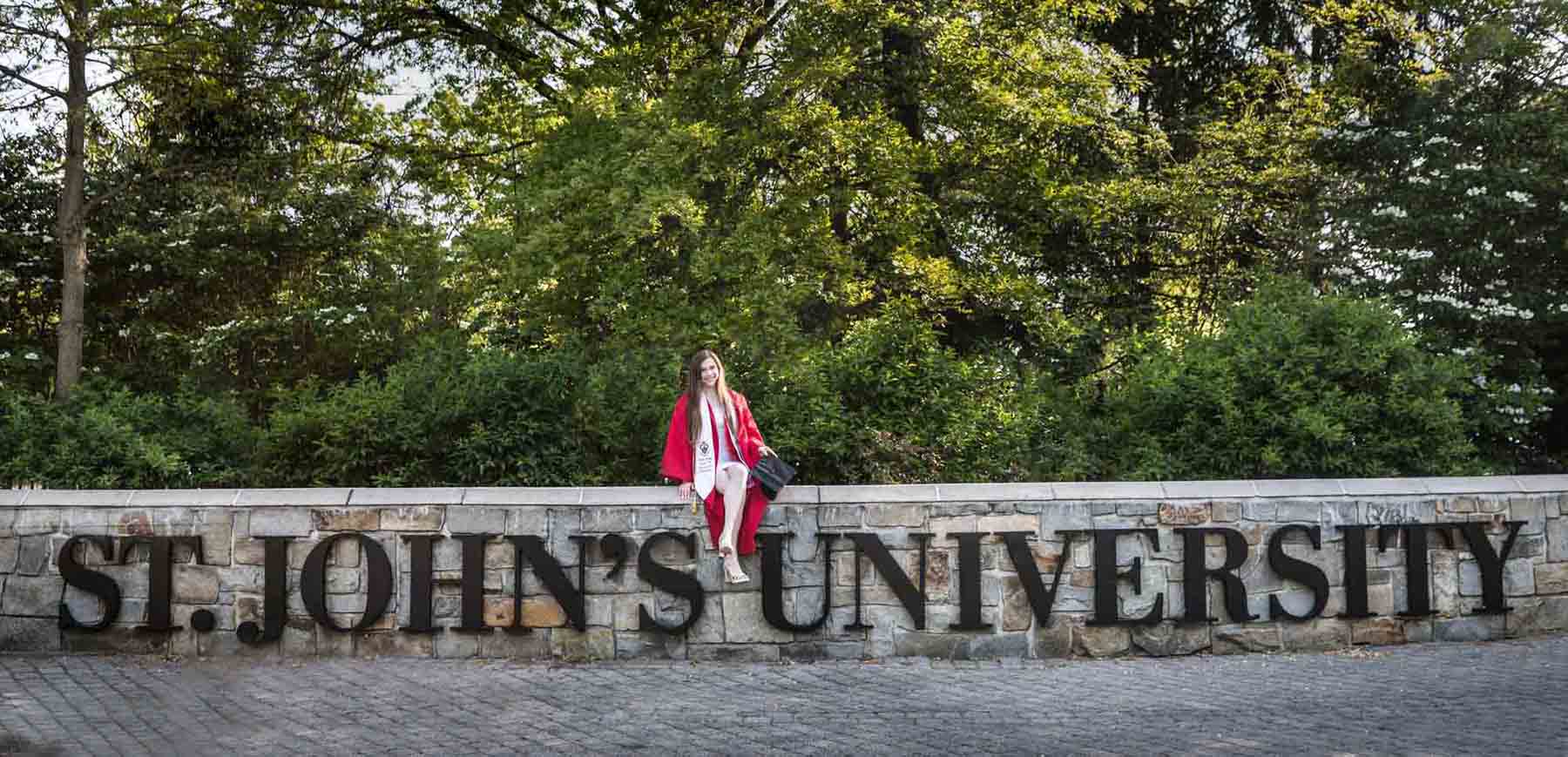 Female graduate wearing red robe and white sashes sitting on sign during a St. John’s University graduate portrait session