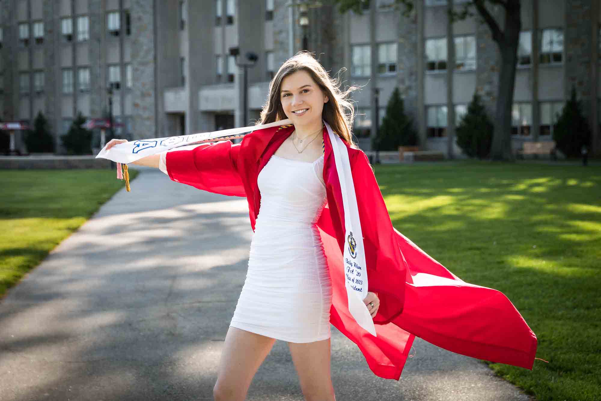 Female graduate on sidewalk wearing red robe and holding out white sashes for an article on graduation portrait photo tips