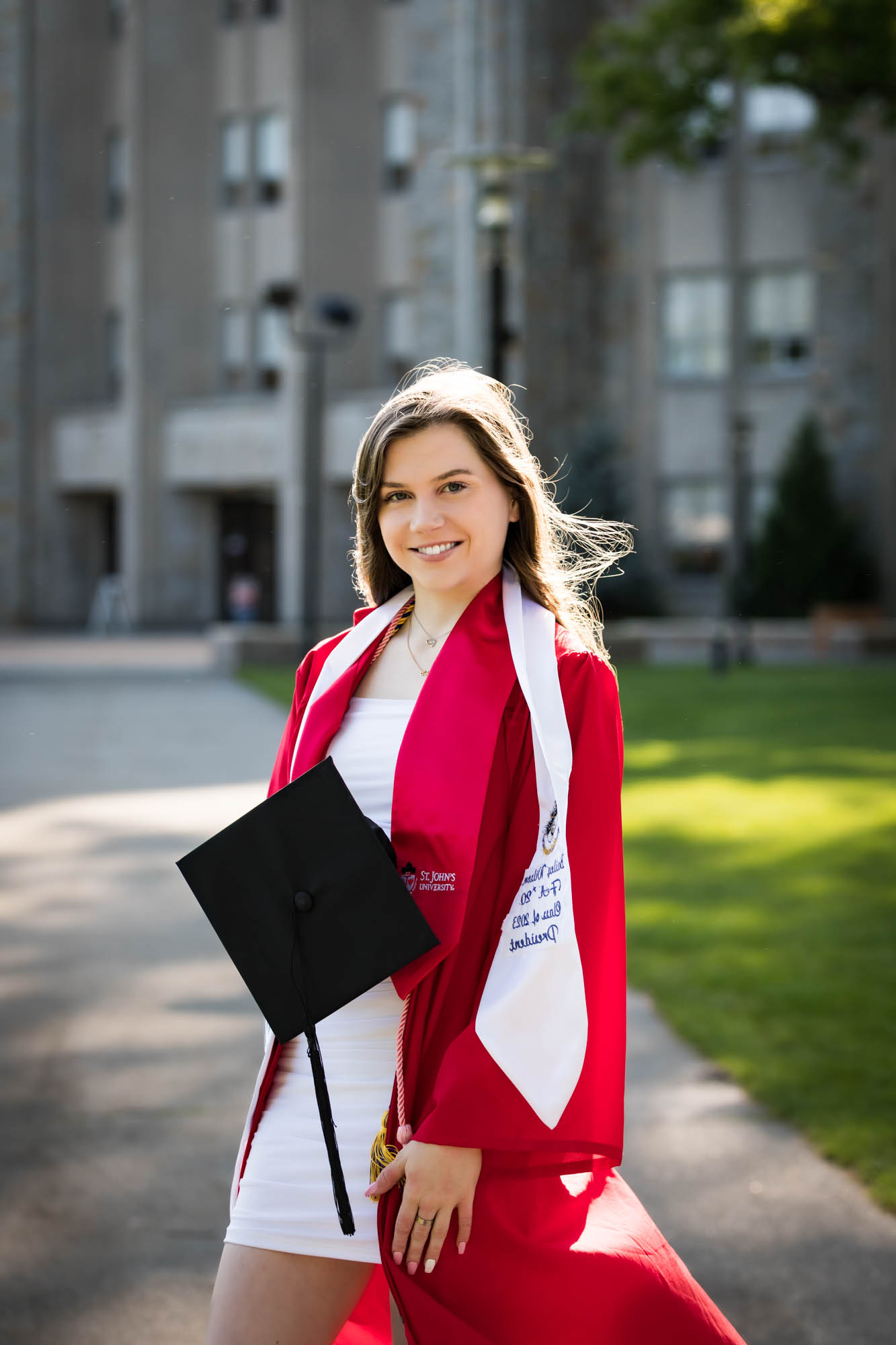 Female graduate wearing red robe and white sash holding cap during a St. John’s University graduate portrait session