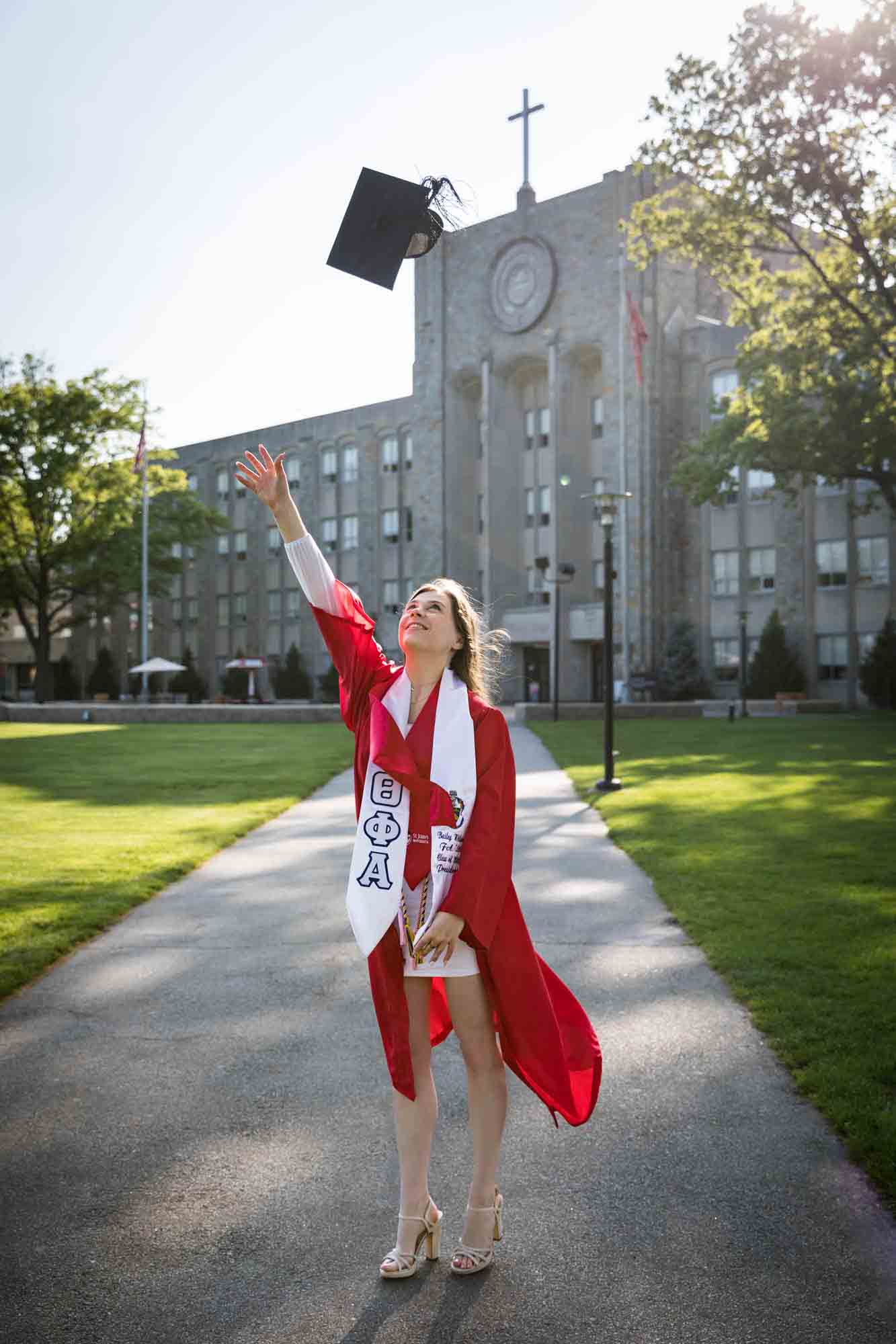 Female graduate wearing red robe and white sash throwing cap in air during a St. John’s University graduate portrait session