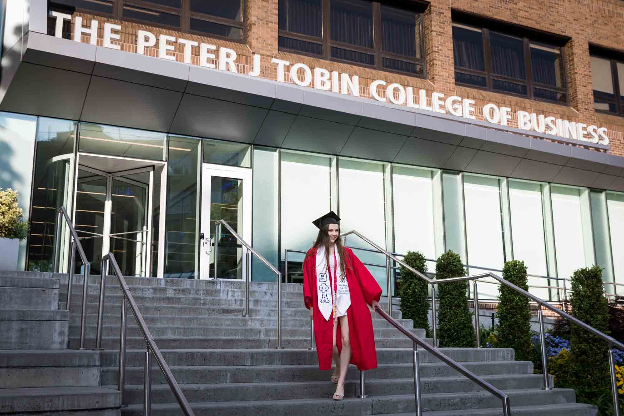 Female graduate wearing red robe and and cap in front of business school during a St. John’s University graduate portrait session