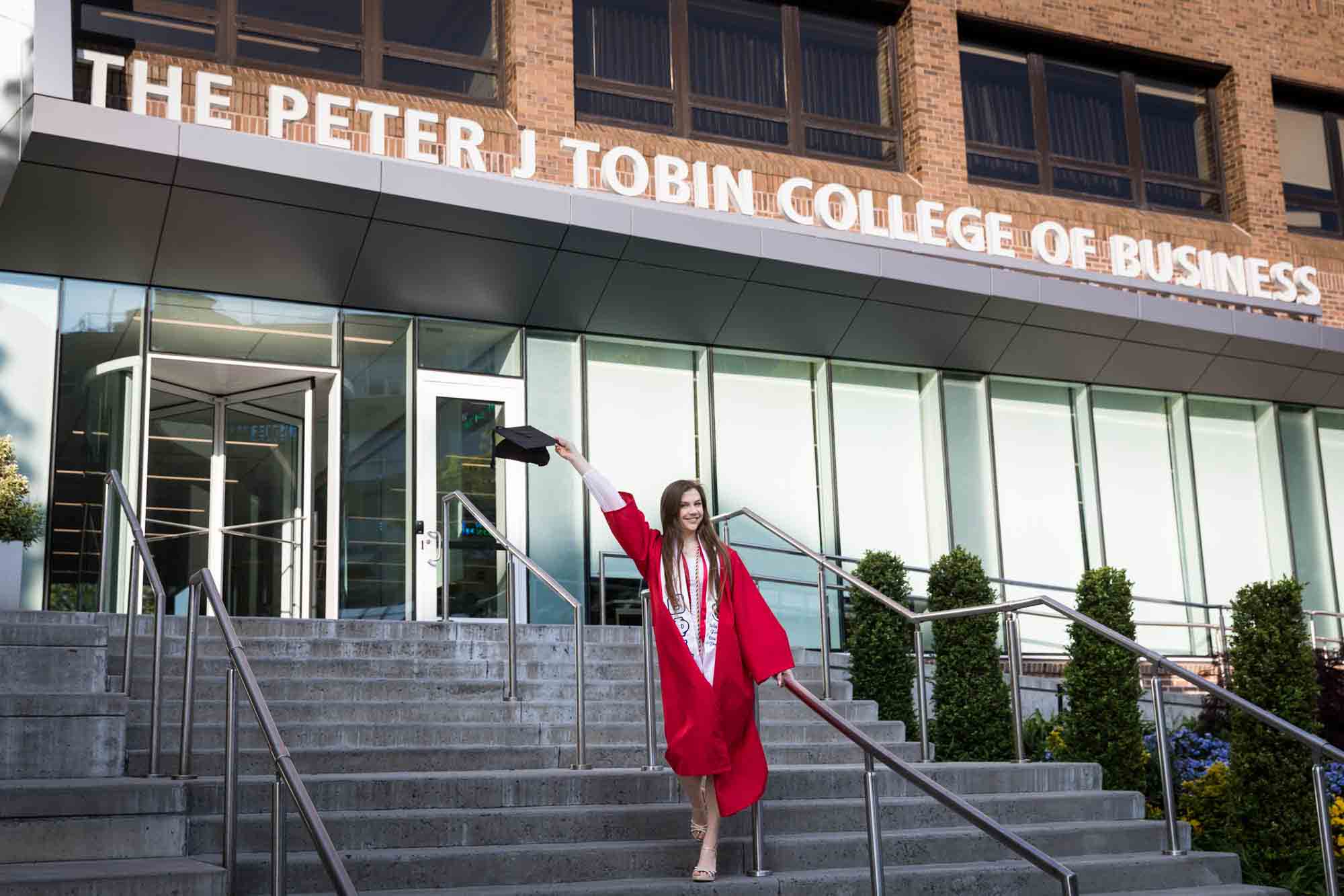 Female graduate wearing red robe and white sash holding cap in air in front of business school during a St. John’s University graduate portrait session