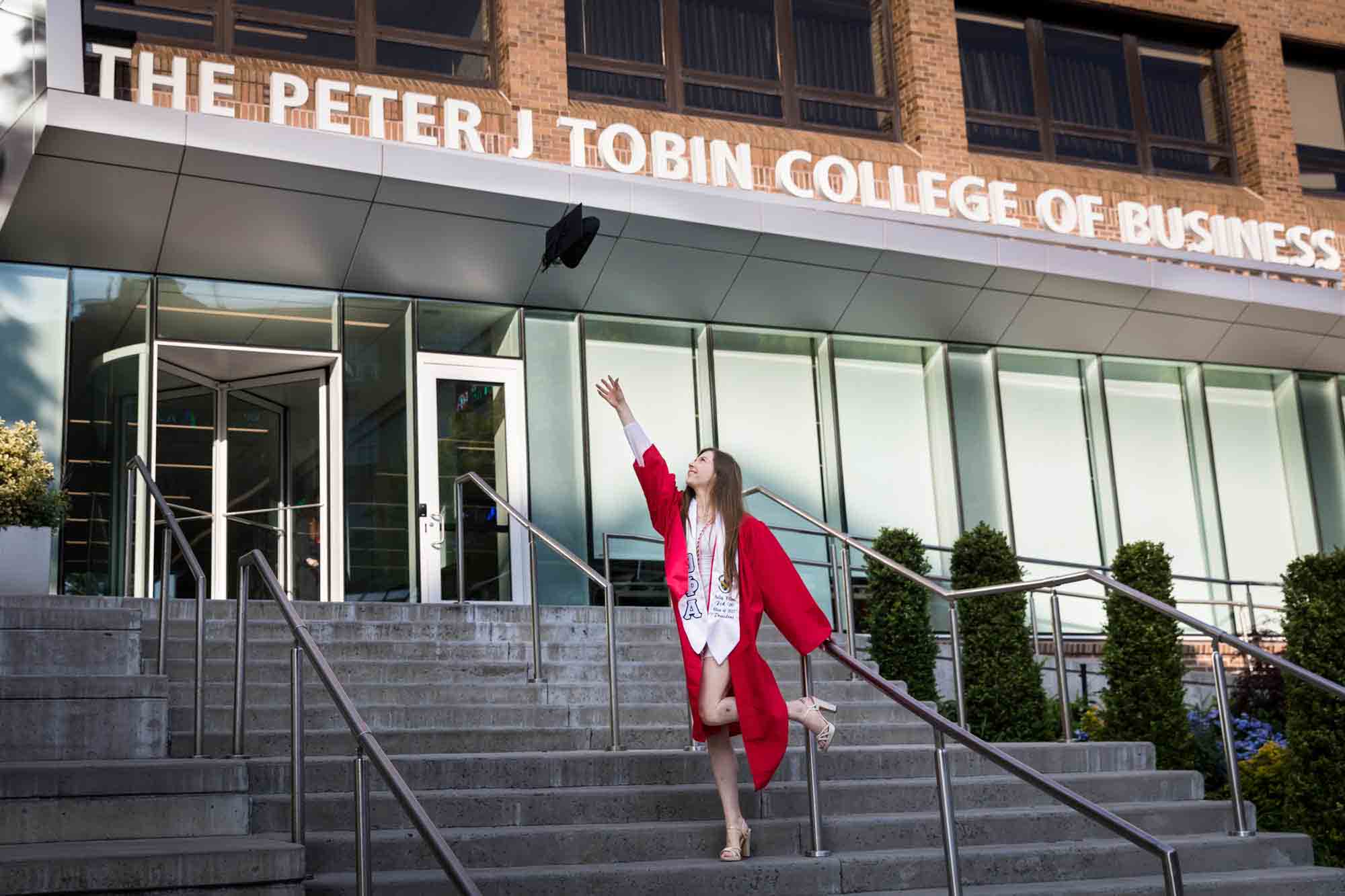 Female graduate wearing red robe and throwing cap in the air on steps of business school for an article on graduation portrait photo tips