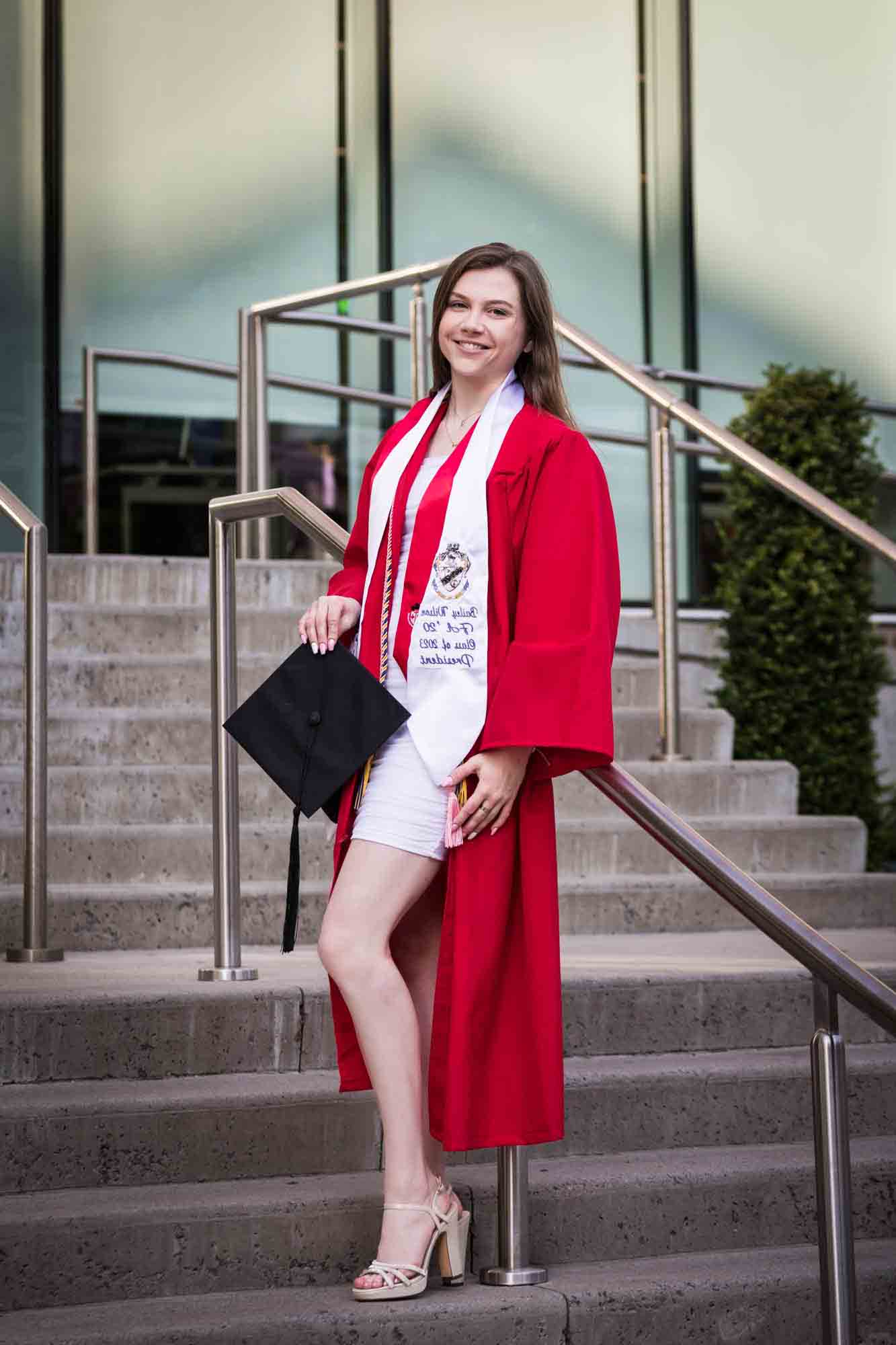 Female graduate wearing red robe and white sash holding cap on steps during a St. John’s University graduate portrait session