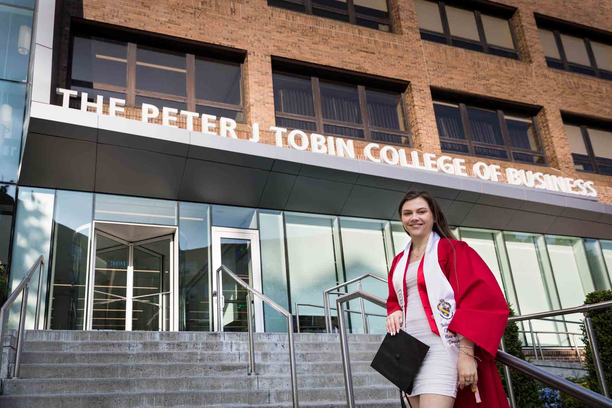 Female graduate wearing red robe and white sash holding cap in front of business school during a St. John’s University graduate portrait session