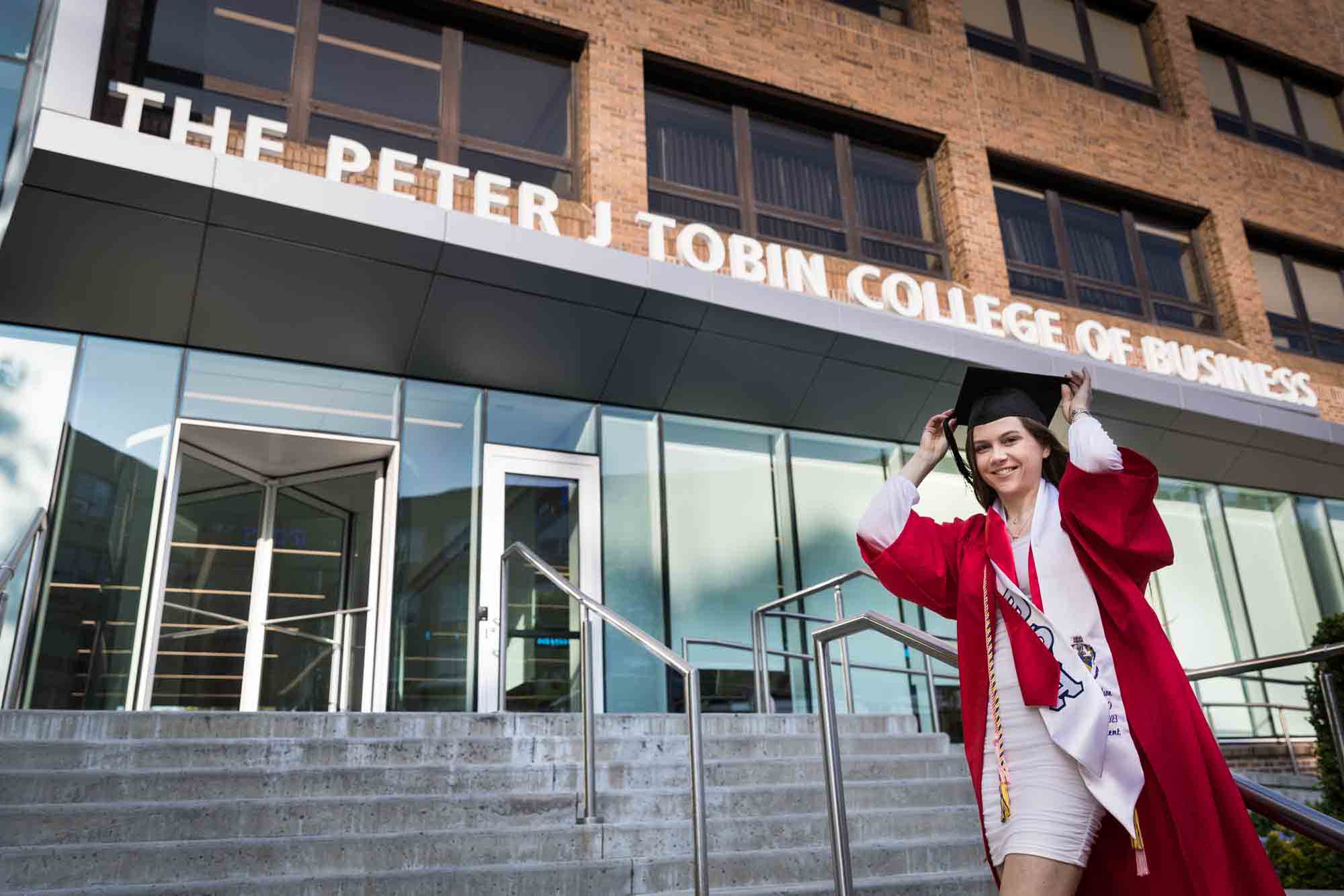 Female graduate wearing red robe and white sash holding cap in front of business school during a St. John’s University graduate portrait session