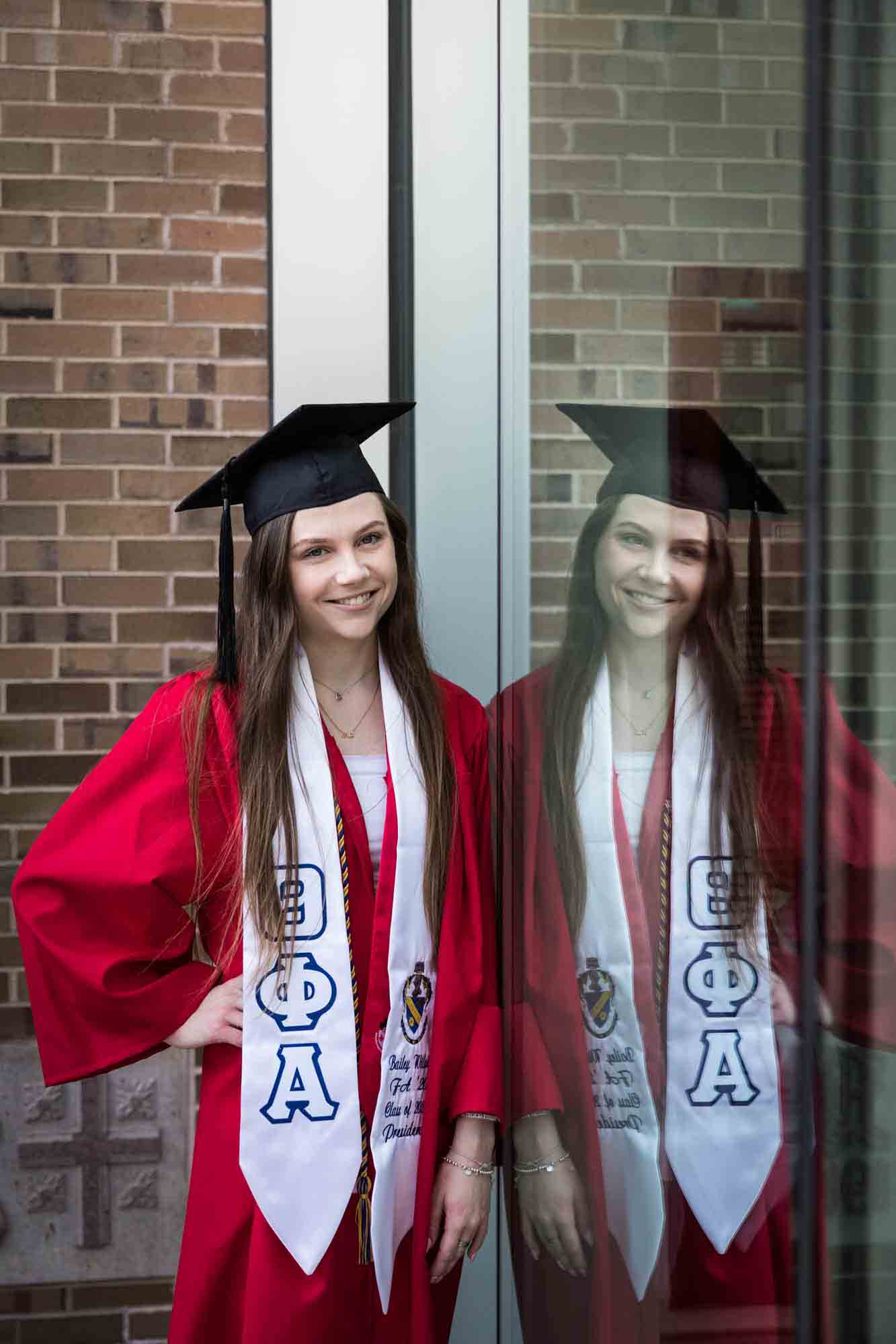Female graduate wearing red robe, cap and white sash leaning against window with reflection during a St. John’s University graduate portrait session