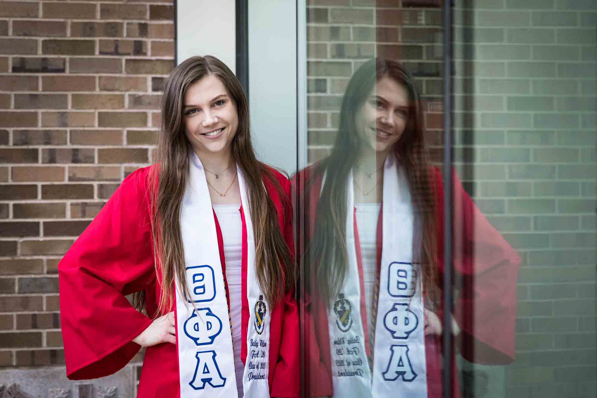 Female graduate wearing red robe and white sash leaning against window with reflection during a St. John’s University graduate portrait session