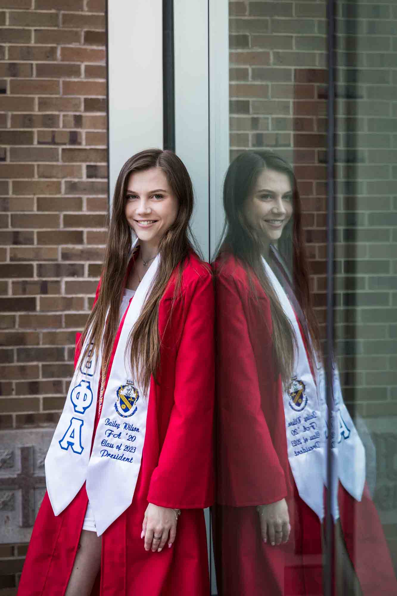 Female graduate wearing red robe and white sash leaning against window with reflection during a St. John’s University graduate portrait session