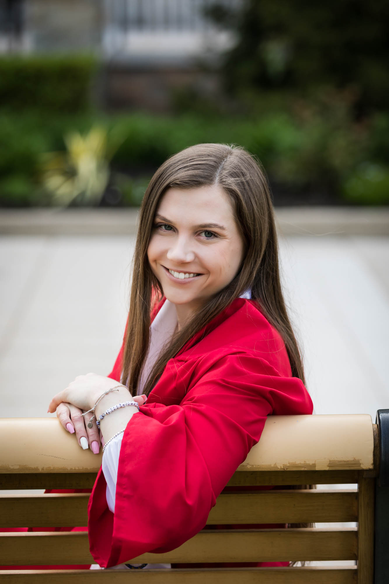 Female grad wearing red rob sitting on bench looking backwards during a St. John’s University graduate portrait session