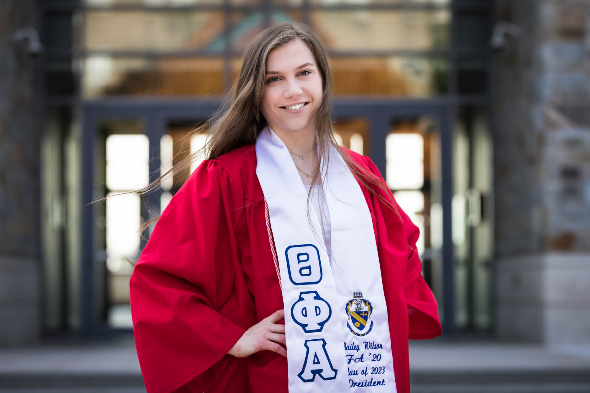 Female graduate wearing red robe and white sash with hands on hips for an article on graduation portrait photo tips