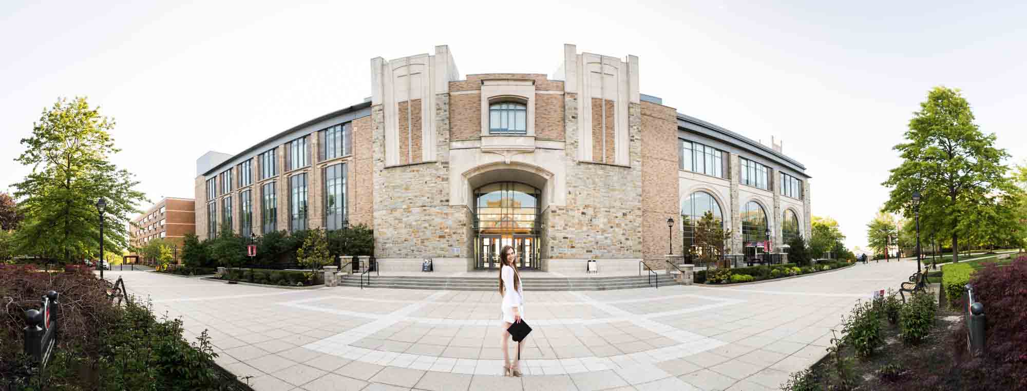 Panorama photo of female graduate wearing white dress and holding cap in front of D'Angelo Building during a St. John’s University graduate portrait session