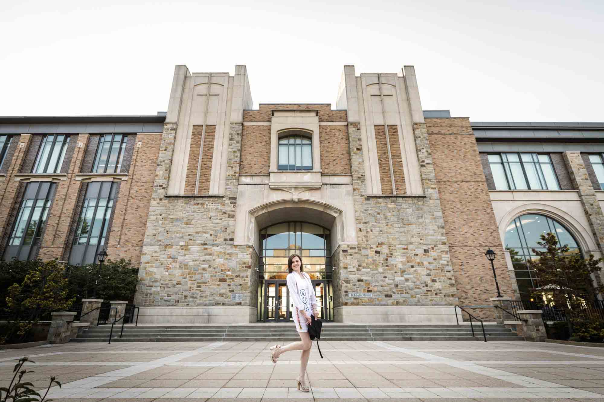 Female graduate wearing white dress and holding cap in front of D'Angelo Building during a St. John’s University graduate portrait session