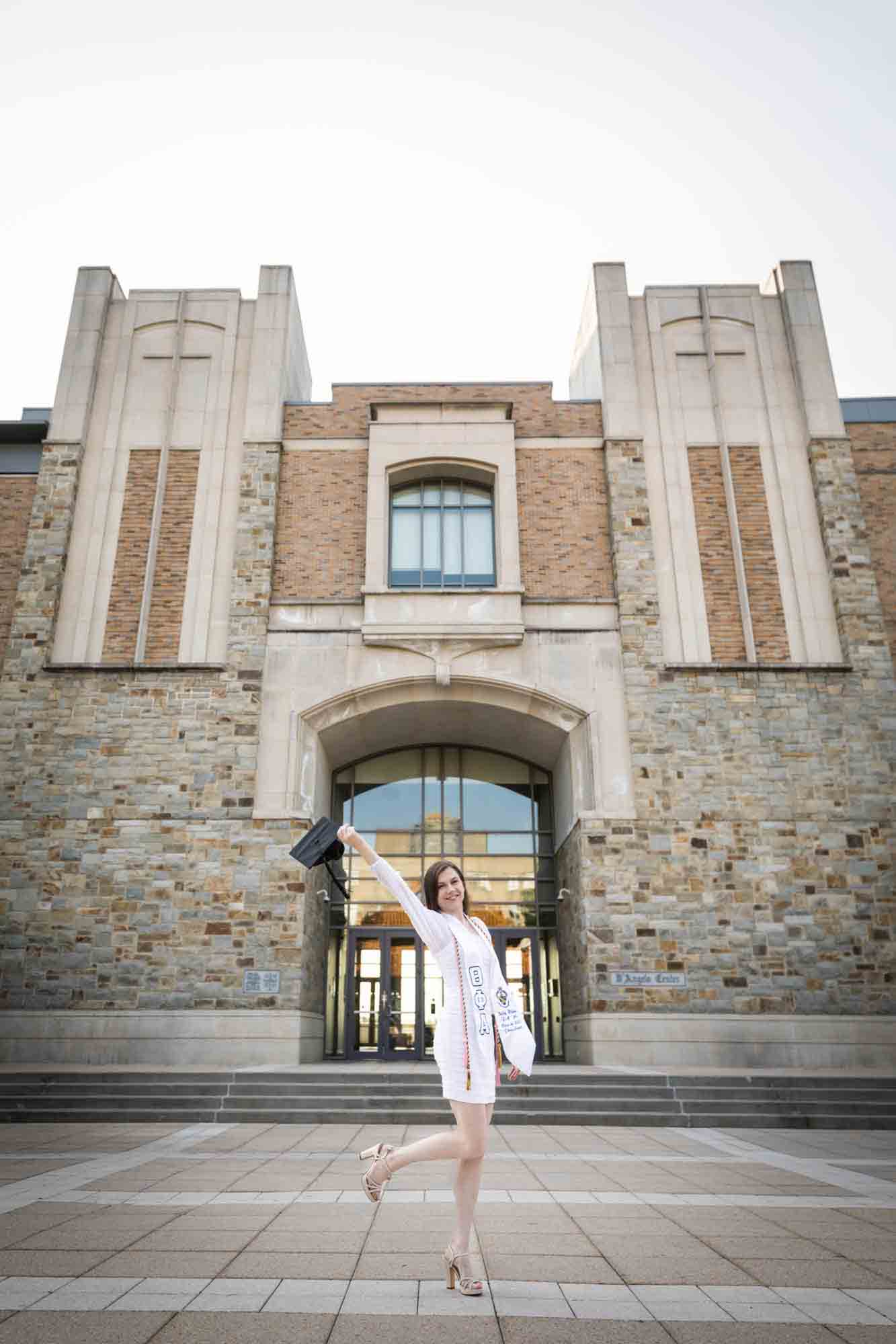 Female graduate wearing white dress and holding cap in the air during a St. John’s University graduate portrait session