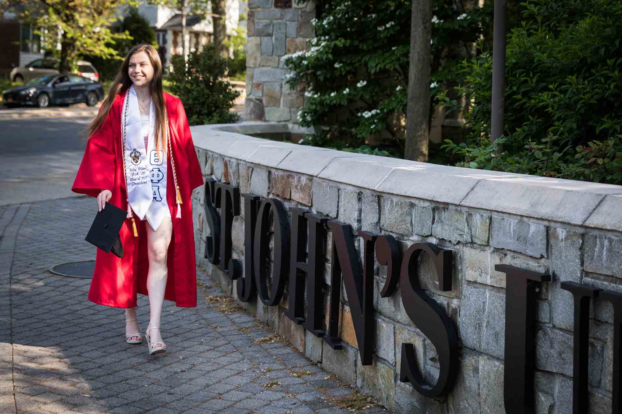 Female graduate wearing red robe and holding cap walking in front of university sign for an article on graduation portrait photo tips