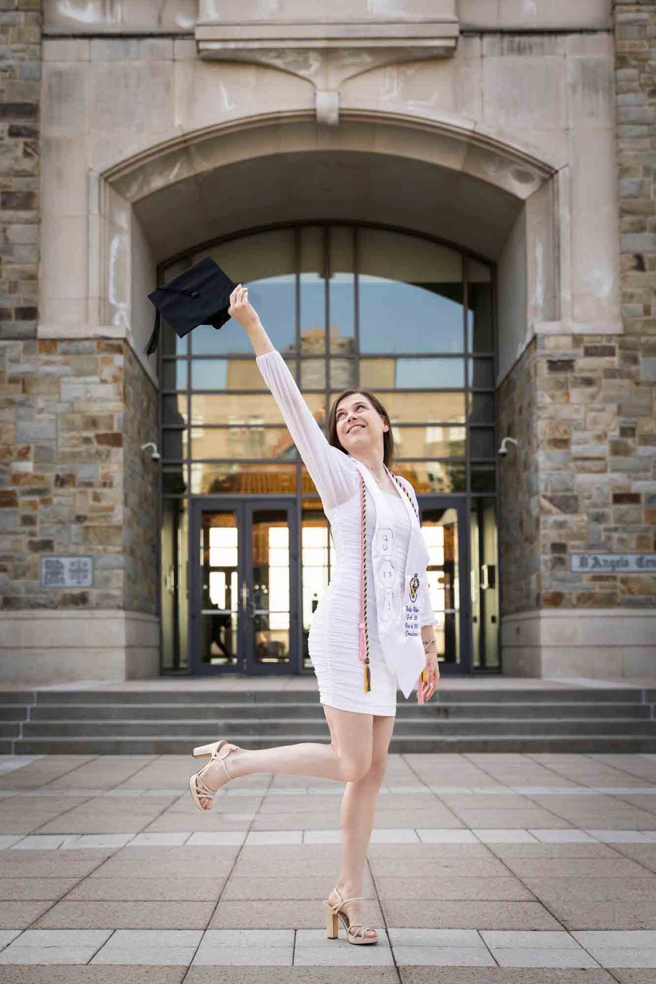 Female graduate wearing white dress and holding cap in the air during a St. John’s University graduate portrait session