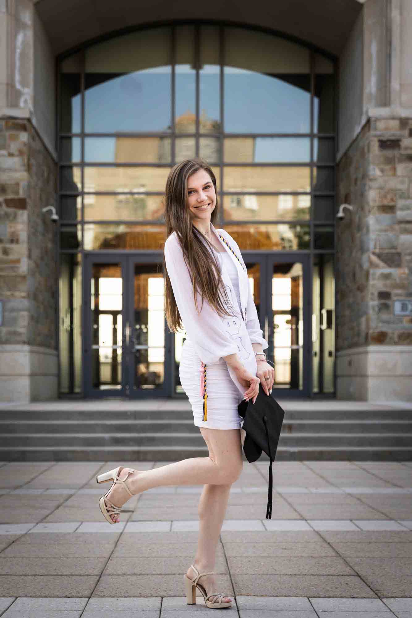 Female graduate wearing white dress and holding cap with leg up during a St. John’s University graduate portrait session