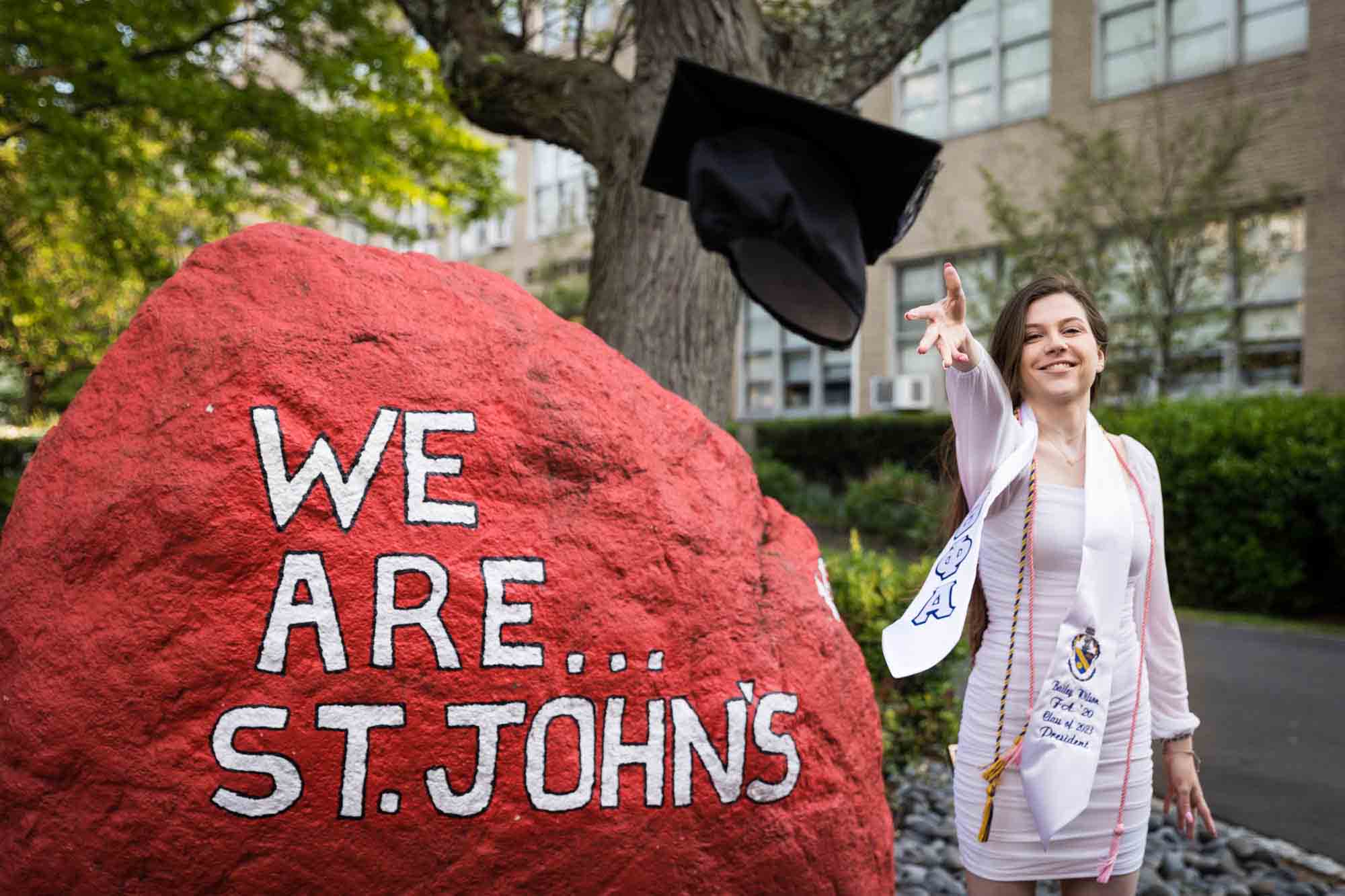 Female graduate wearing white dress standing by spirit rock throwing cap during a St. John’s University graduate portrait session